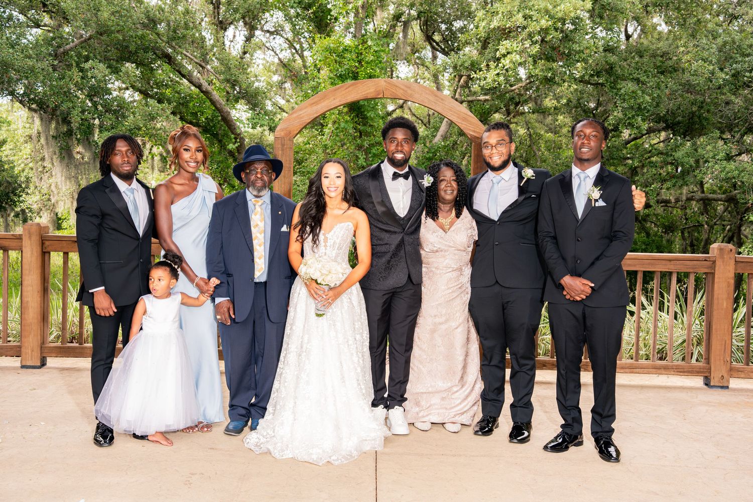 Wedding party group photo under wooden arch surrounded by lush greenery.