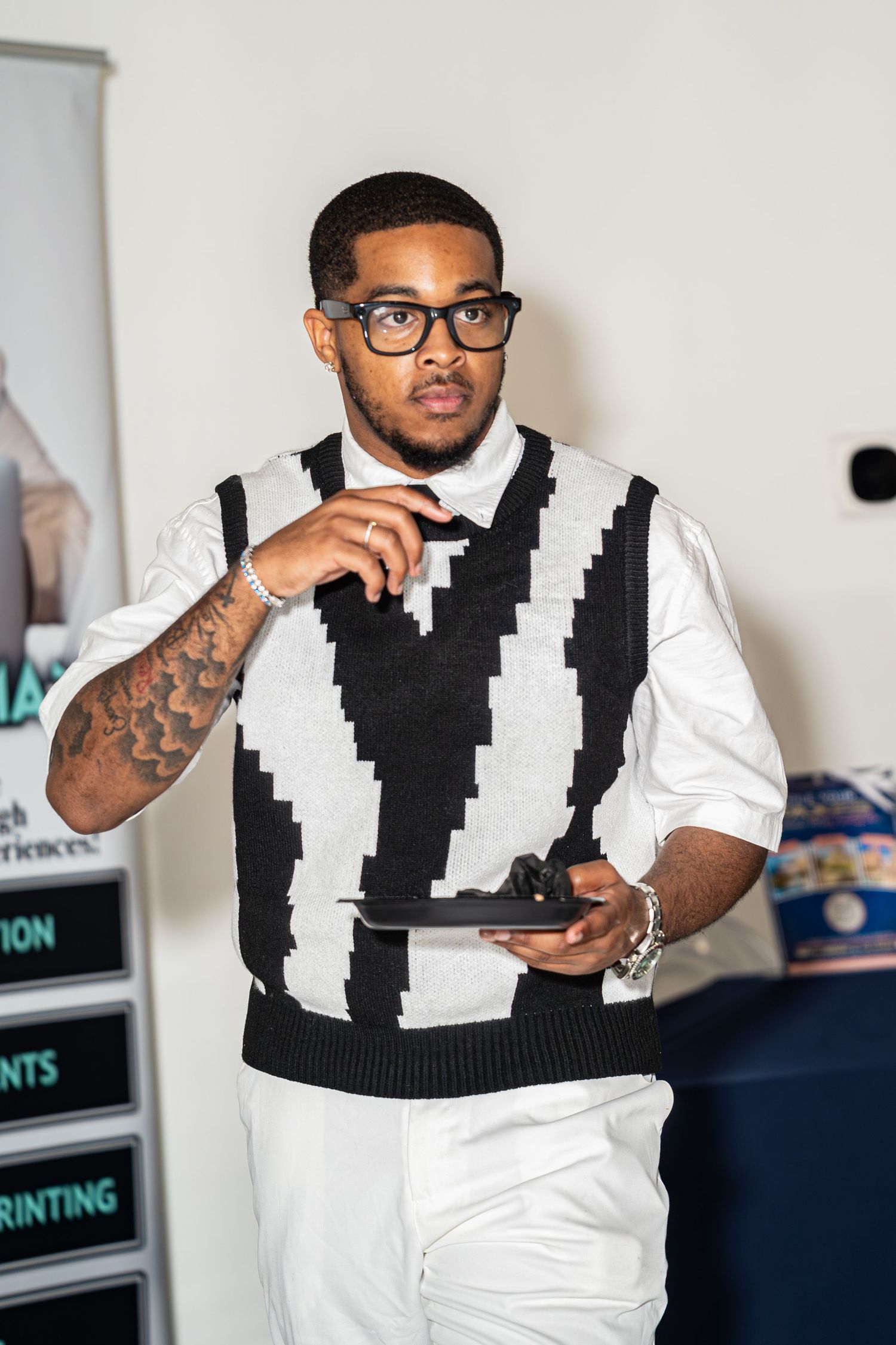 Tattooed man in black and white sweater vest holds tablet at entrepreneur event.