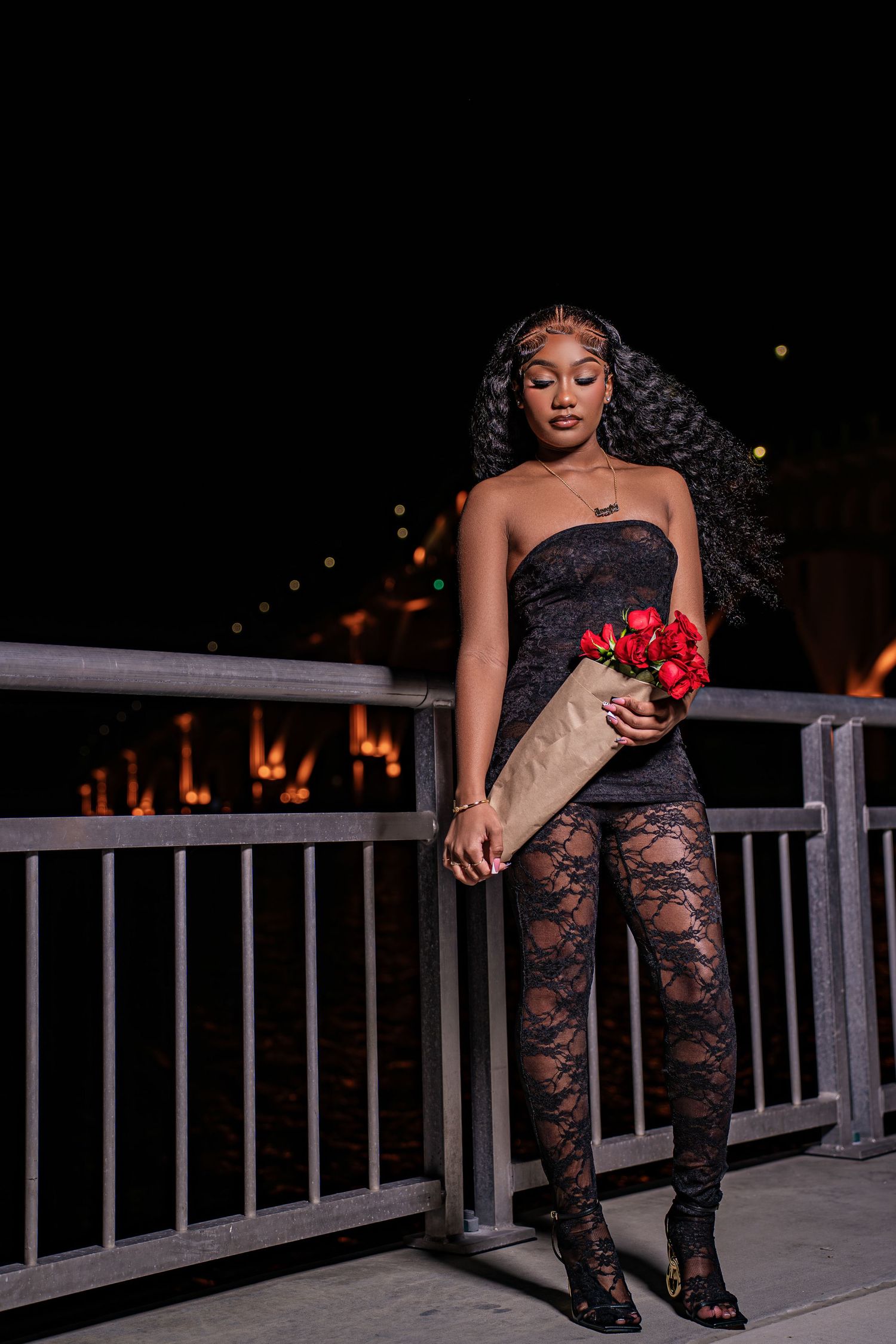 Stylish Black woman in black lace jumpsuit holds red roses bouquet while posing by railing at night.