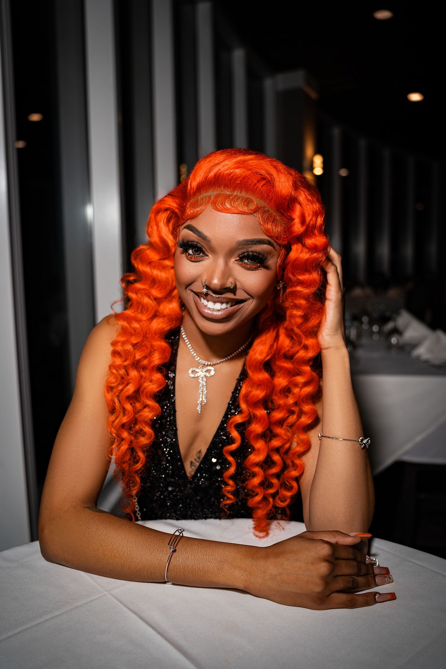 Smiling woman with bright orange curly hair in black dress seated at white tablecloth restaurant table.