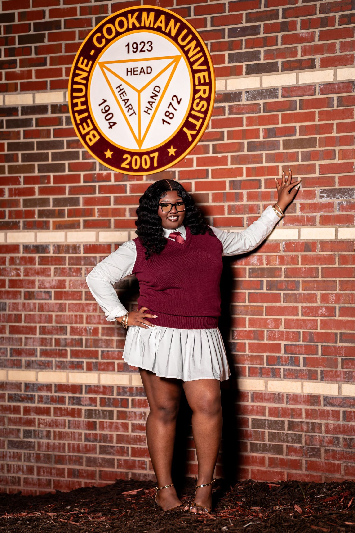 A smiling Black woman in maroon and white outfit poses by Bethune-Cookman University seal on brick wall.