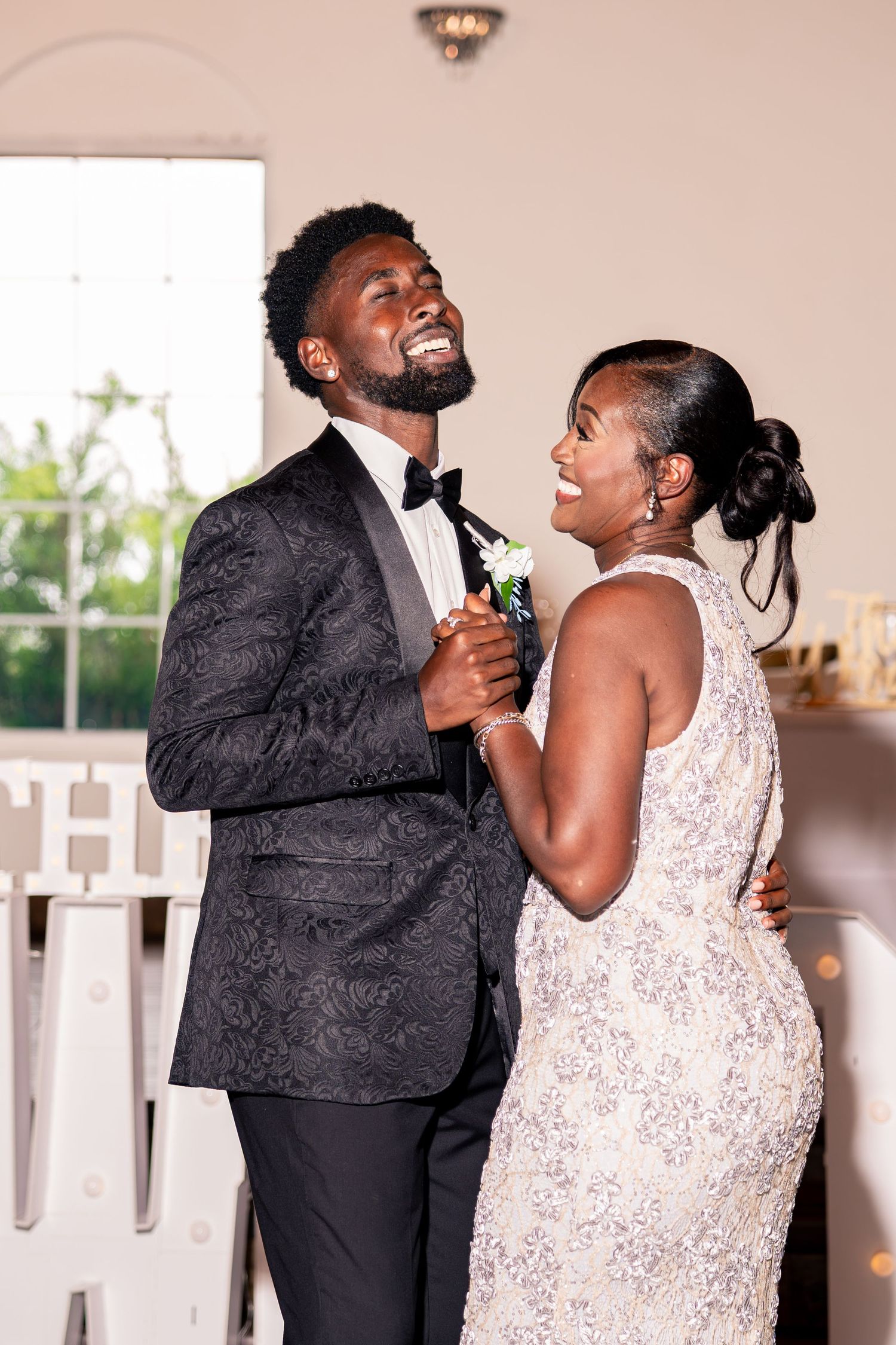 Happy couple shares first dance, groom in black patterned tuxedo and bride in beaded halter wedding dress.