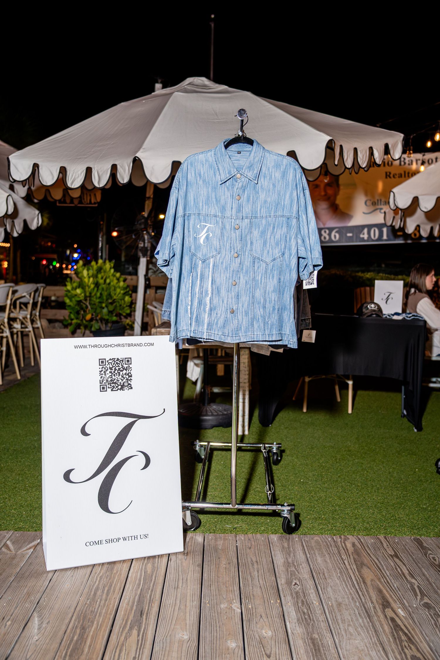 Light blue oversized denim button-up shirt hanging on a rack beside a TC brand sign at an outdoor event.