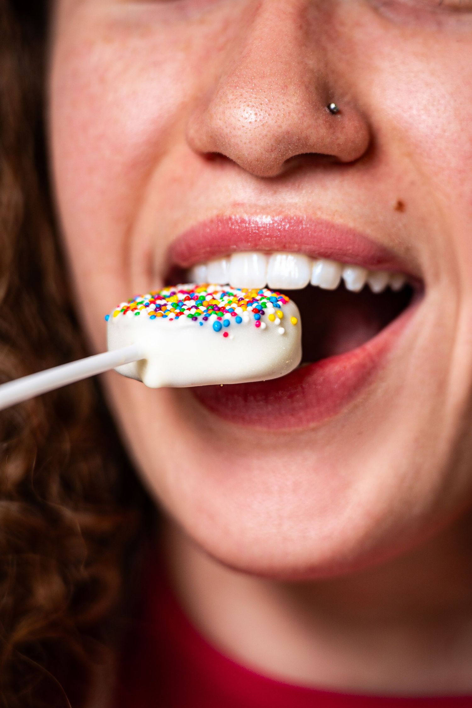Close-up of a smiling woman with a nose piercing biting into a white chocolate sprinkle cake pop.