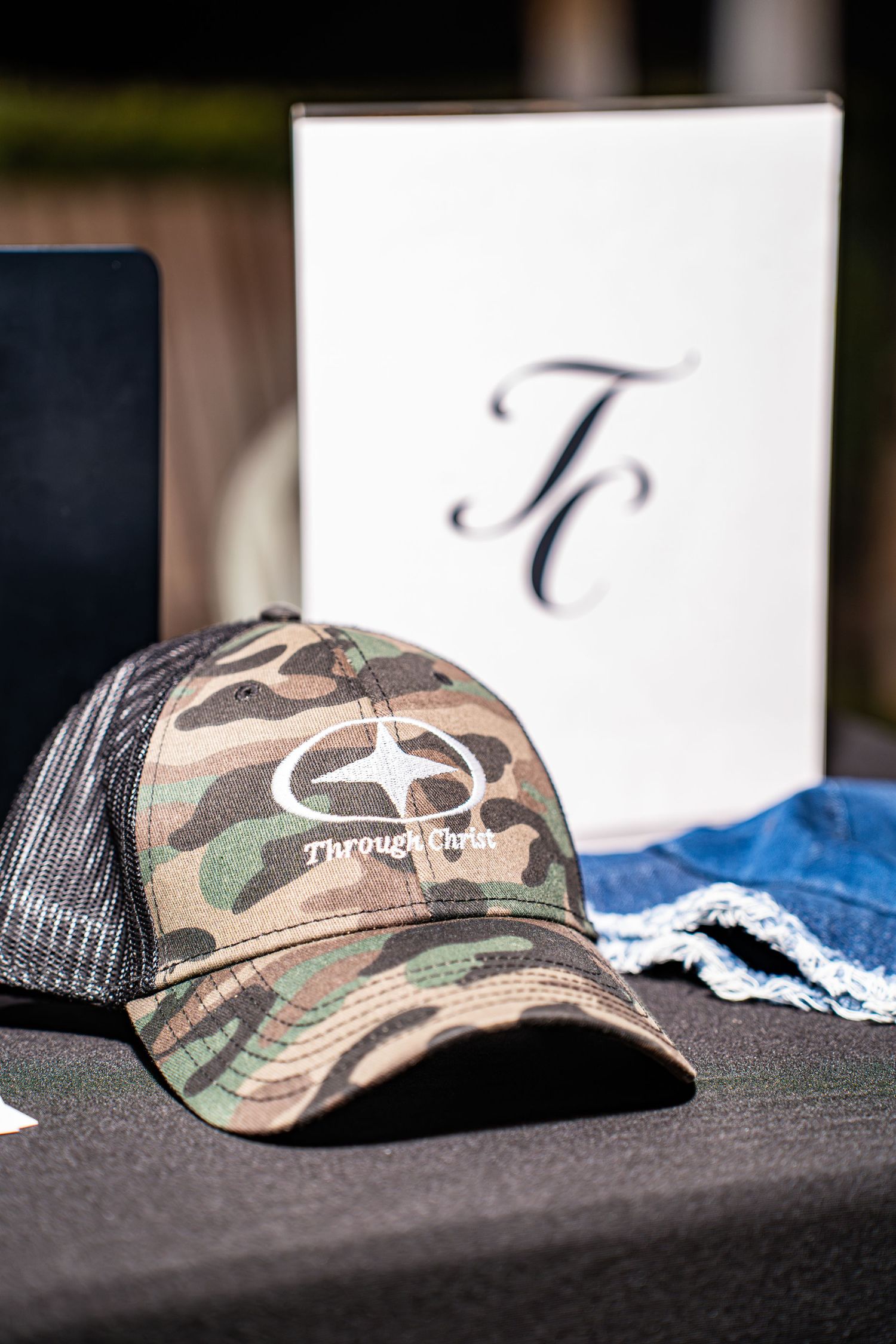 Camouflage Through Christ Brand trucker hat displayed on a vendor table at night.