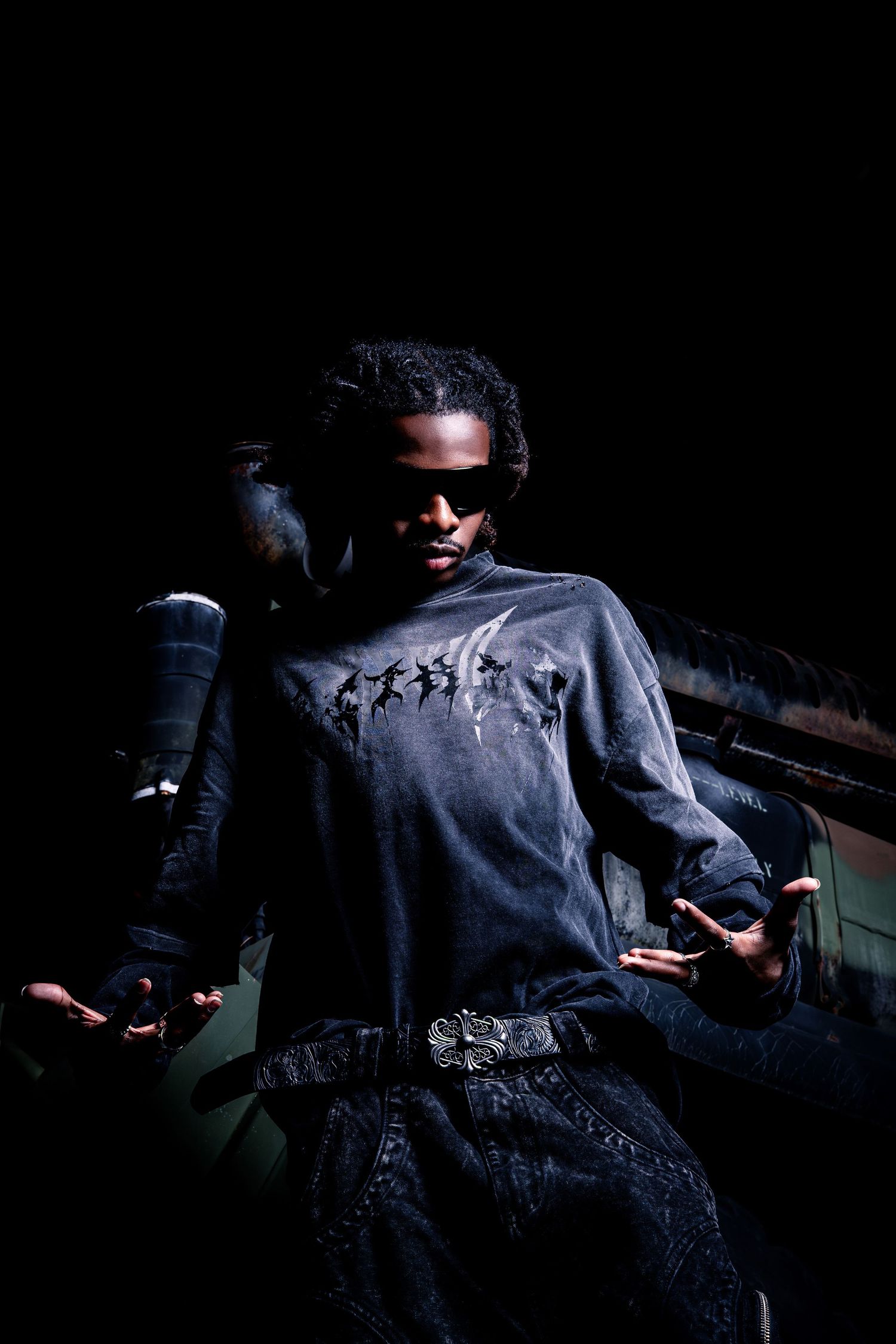 Young Black man in gray long sleeve shirt and sunglasses posing dramatically in dark urban setting.