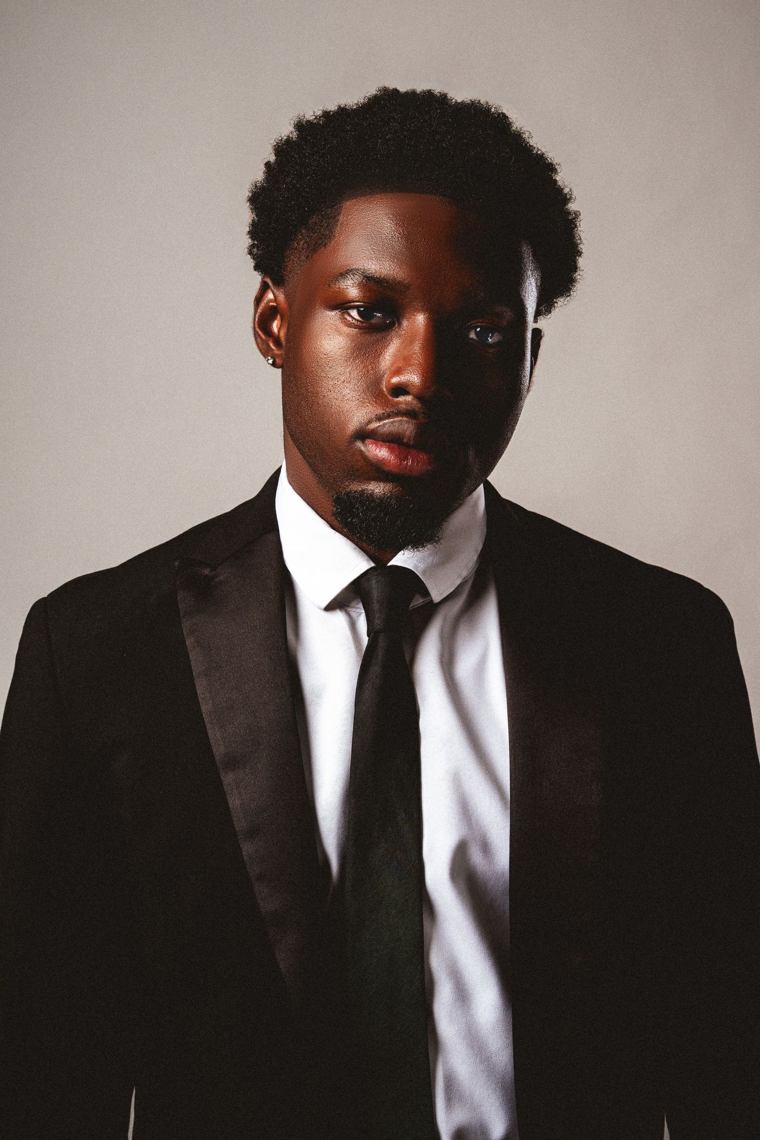 Confident Black man in black tuxedo and tie against gray background.