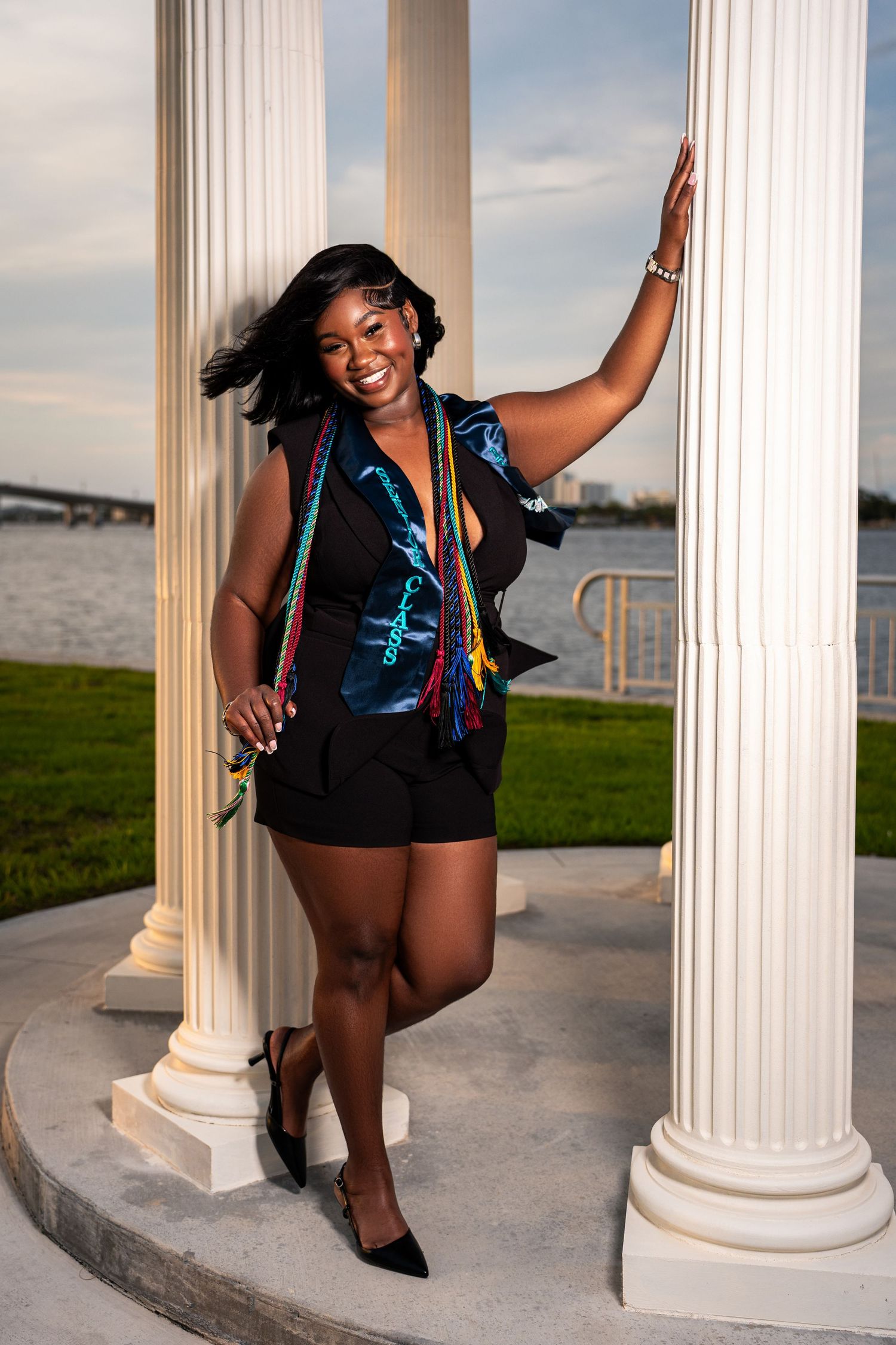 Smiling Black woman in black outfit with colorful graduation stole posing between white columns near river.