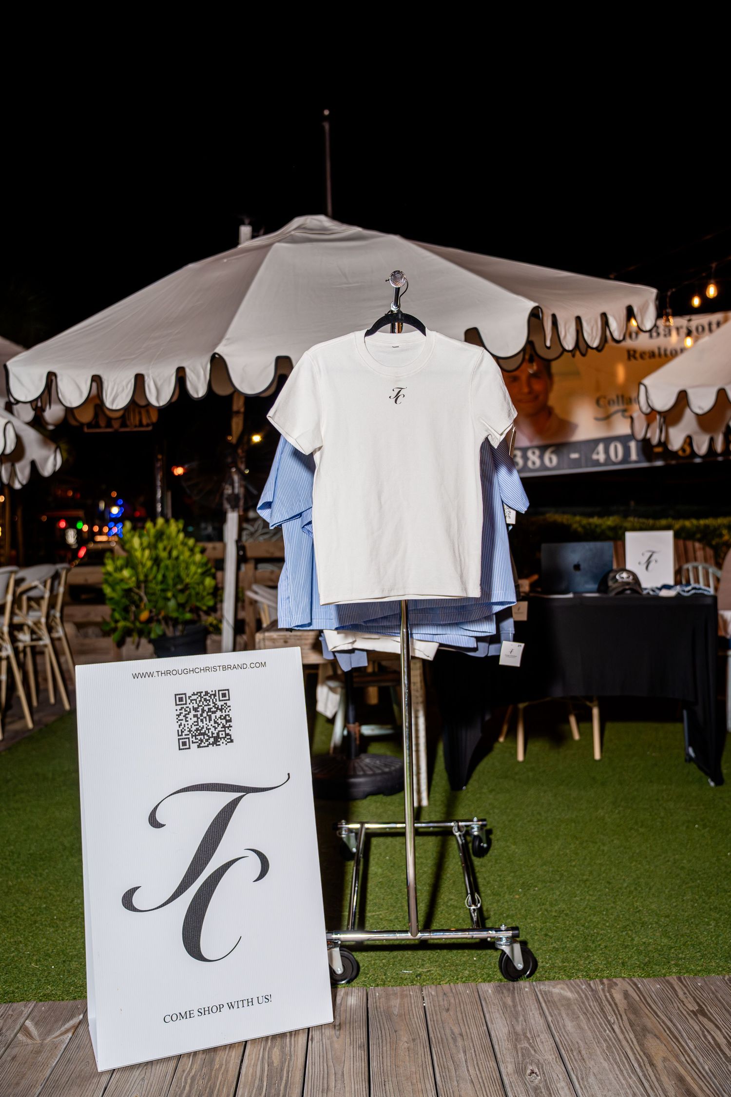 TC brand white t-shirt displayed on rack with QR code sign at outdoor night market.