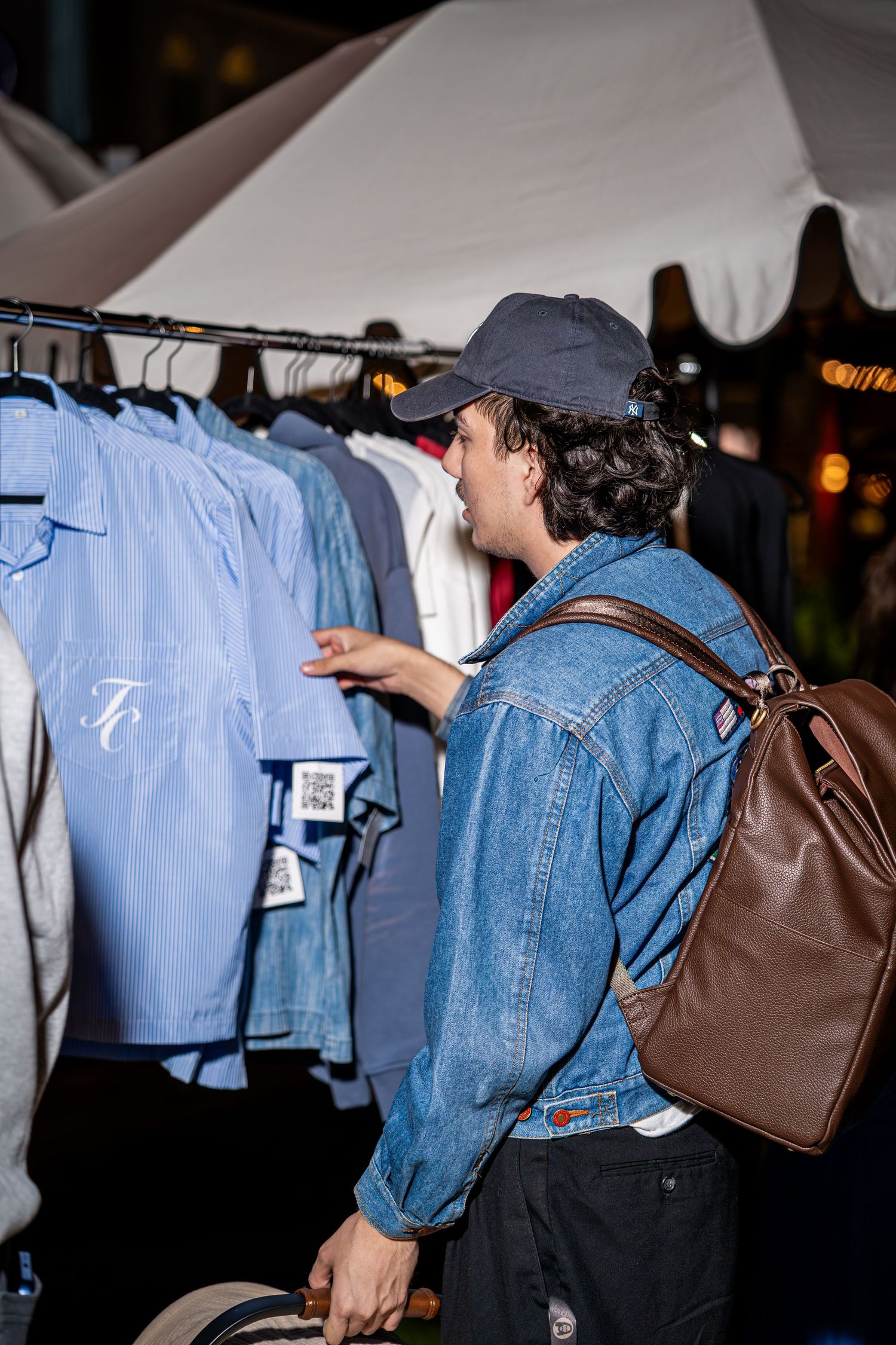 Man in denim jacket and cap browsing blue shirts on rack at outdoor clothing market.
