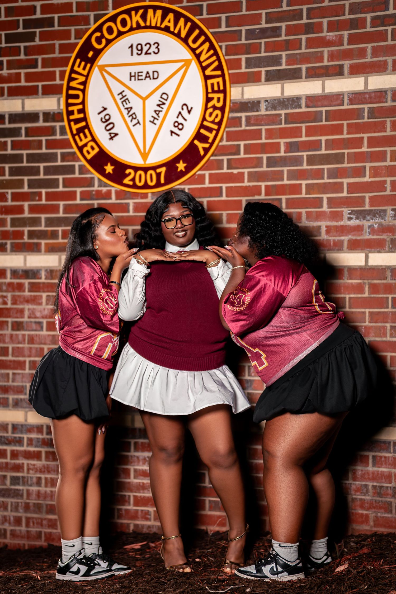 Three young Black women pose together in front of Bethune-Cookman University seal on a brick wall.