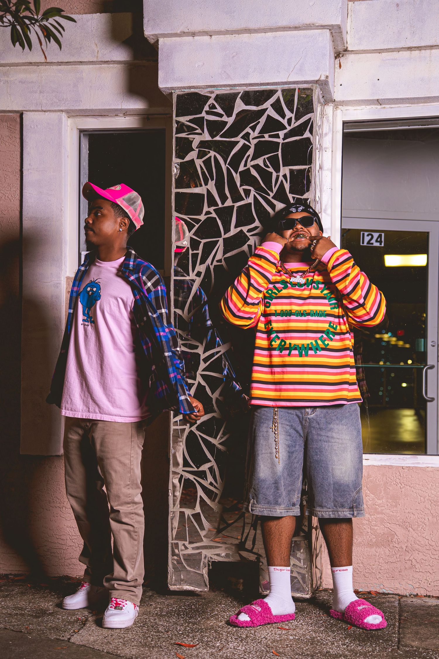 Two stylish young men posing at night outside building wearing colorful streetwear including striped sweater and pink cap.