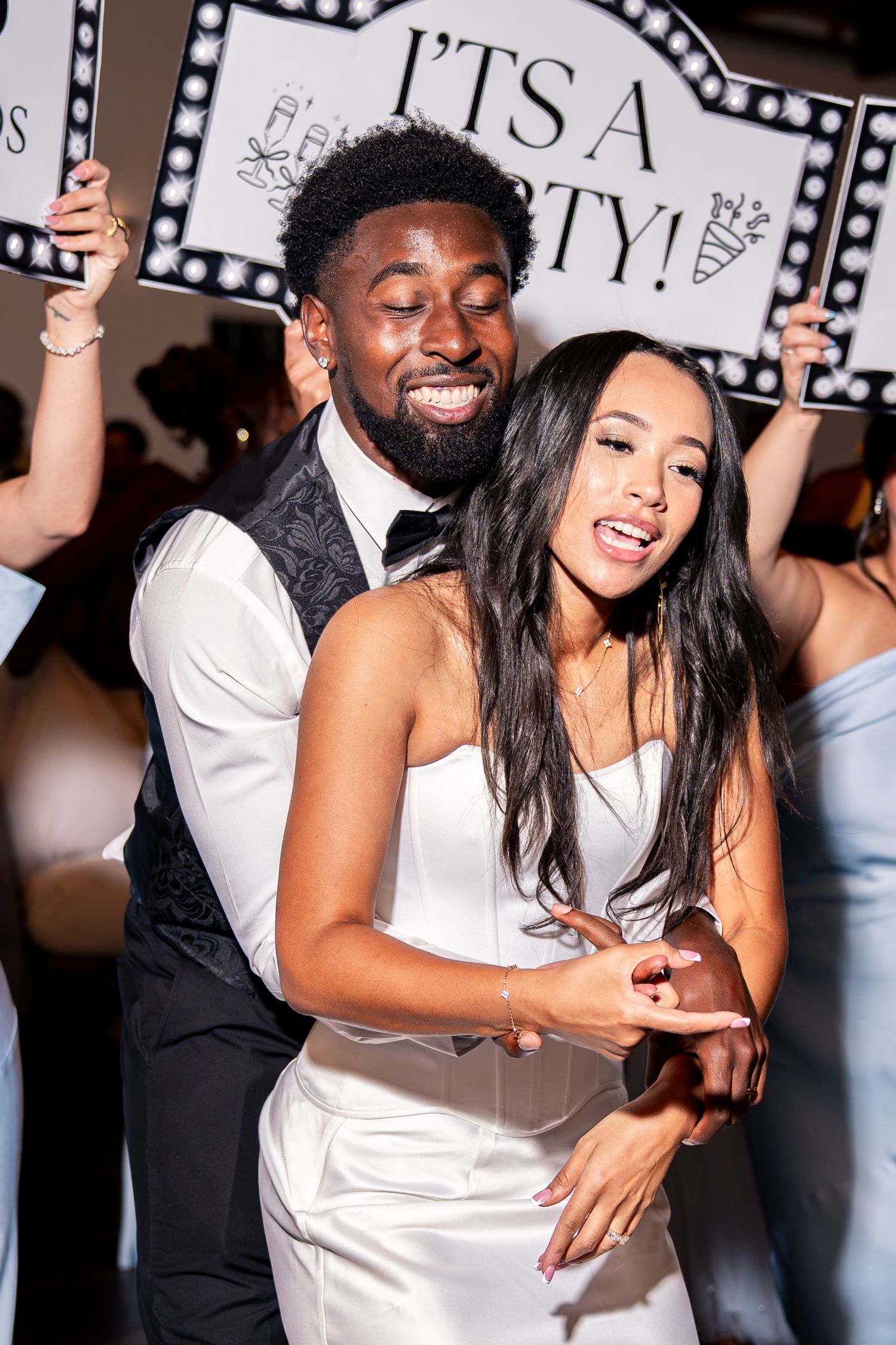 Bride in white dress and groom in vest dance joyfully as guests hold party signs at wedding reception.