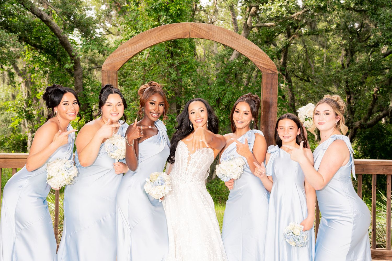 Bride in white lace gown with six bridesmaids in light blue dresses holding bouquets under wooden arch outdoors.