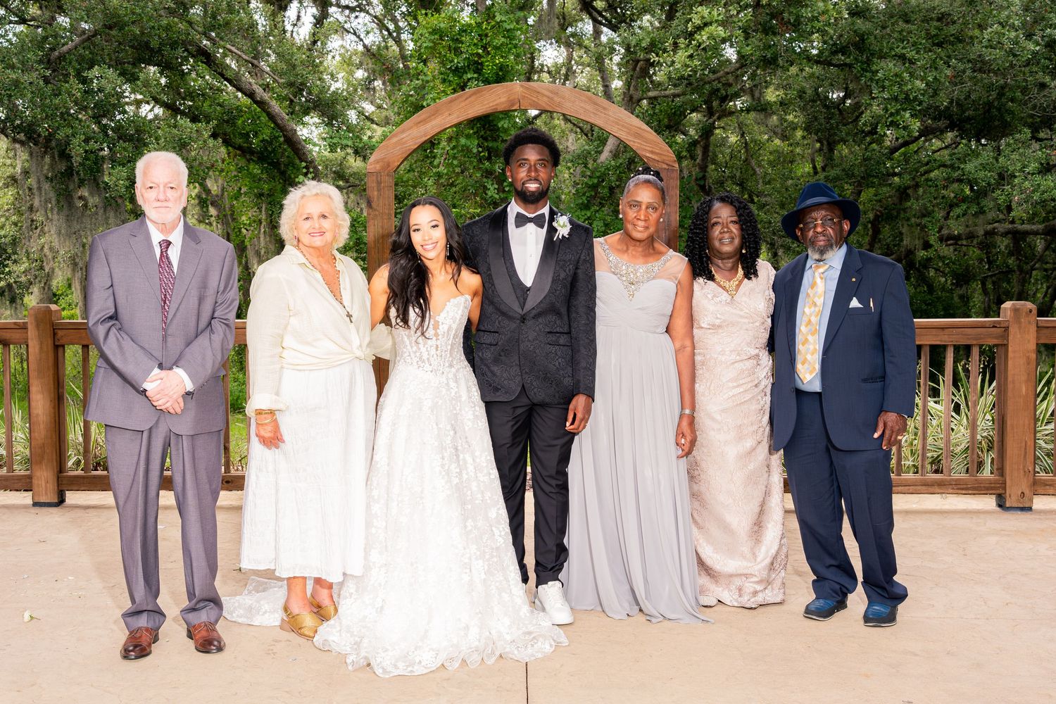 Wedding family portrait of seven people posed under wooden arch at outdoor ceremony.