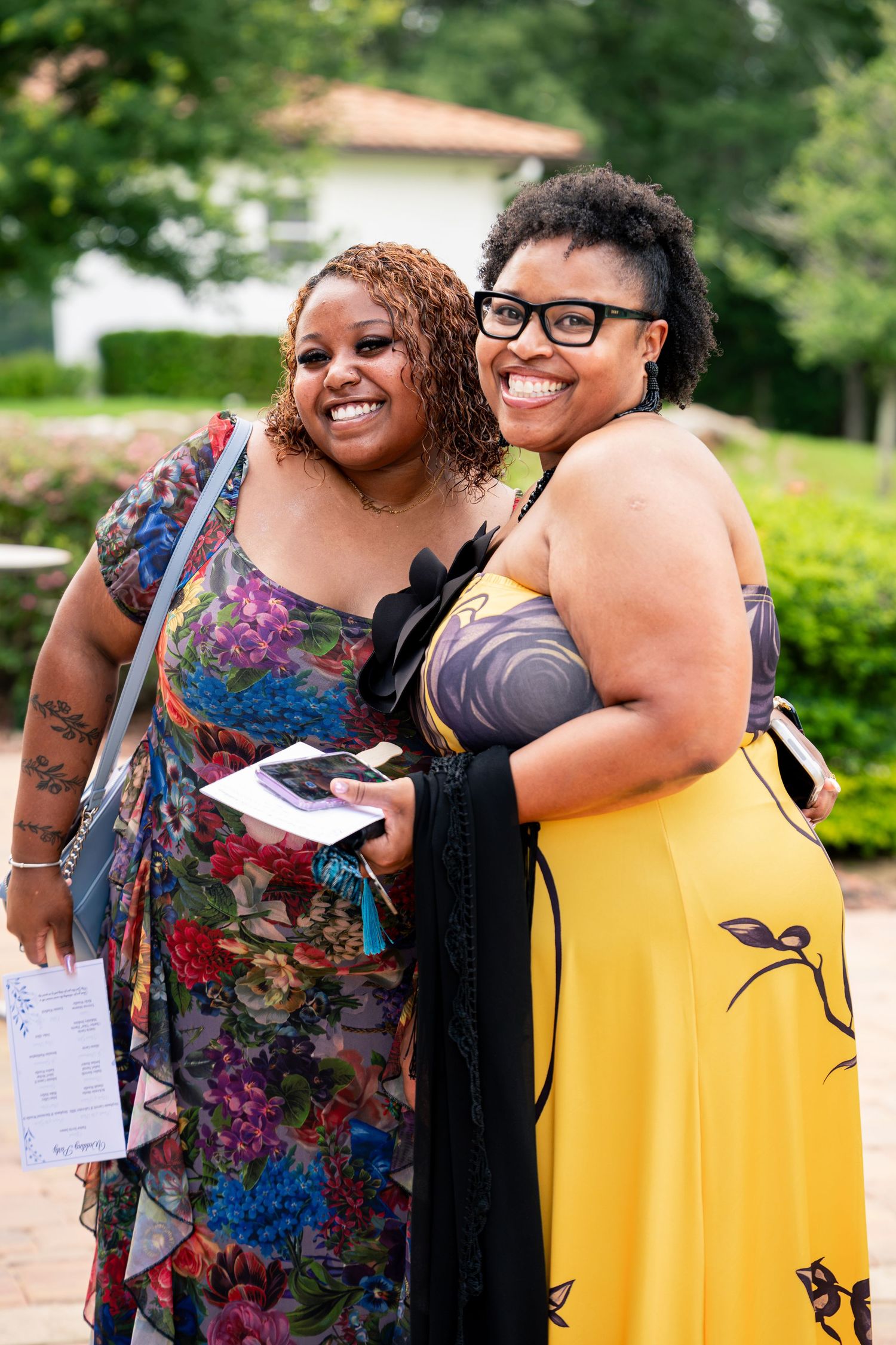 Two smiling women in colorful dresses pose together as wedding guests outdoors in a lush green setting.