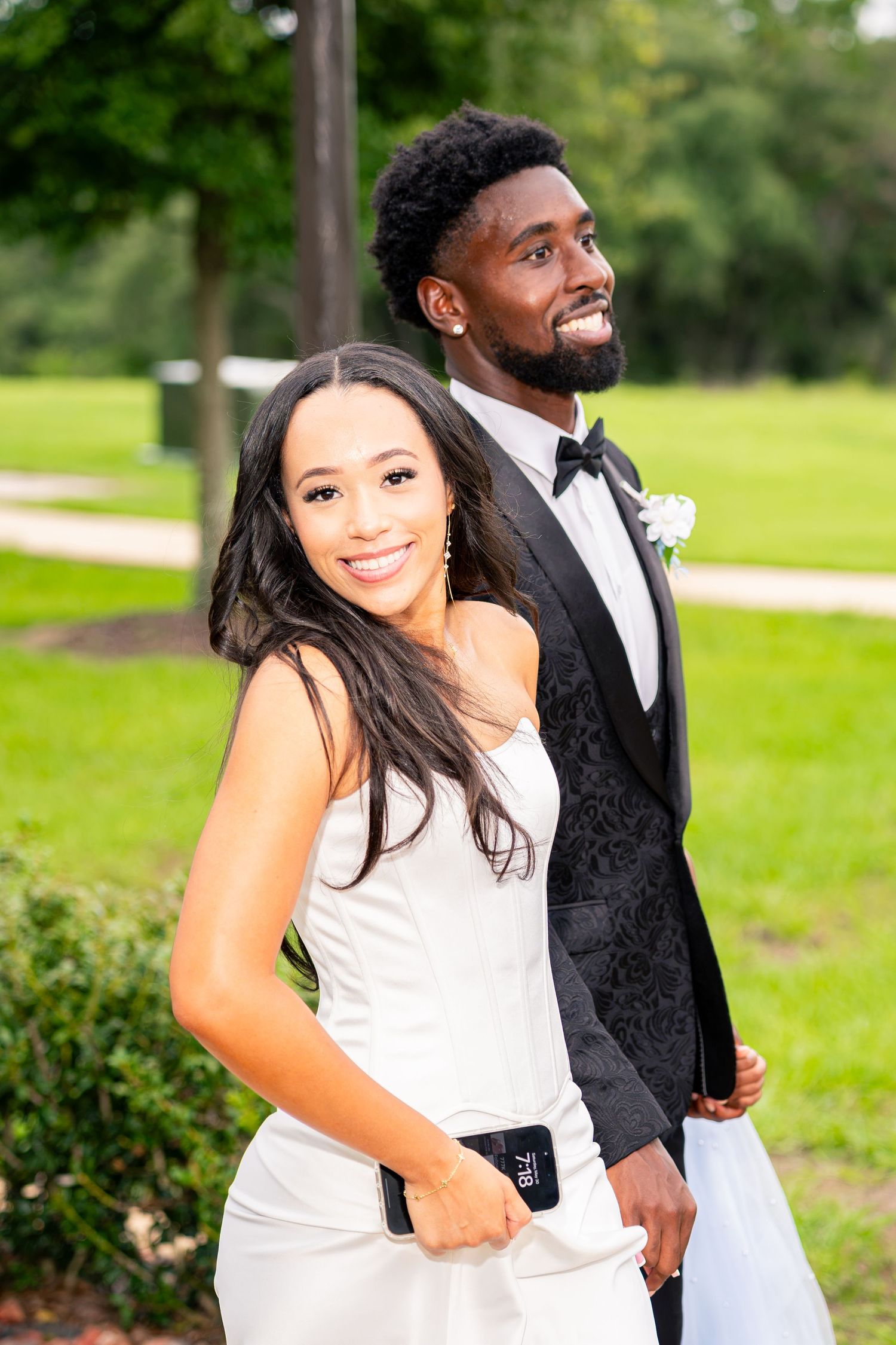 Smiling bride in white gown leans against groom in black patterned tuxedo during outdoor wedding portrait.