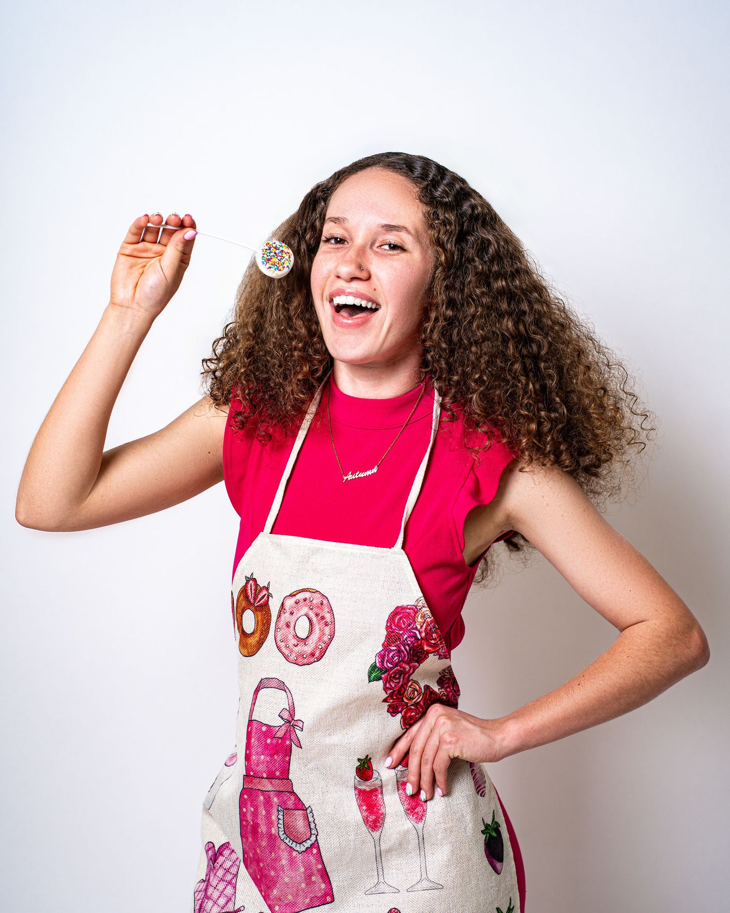 Cheerful young woman in red top and baking apron holding a sprinkle lollipop with curly hair.