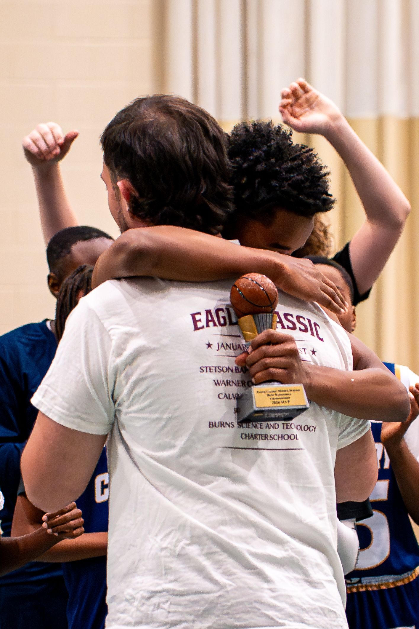 Basketball player holding trophy receives celebratory hug at Eagle Classic tournament award ceremony.
