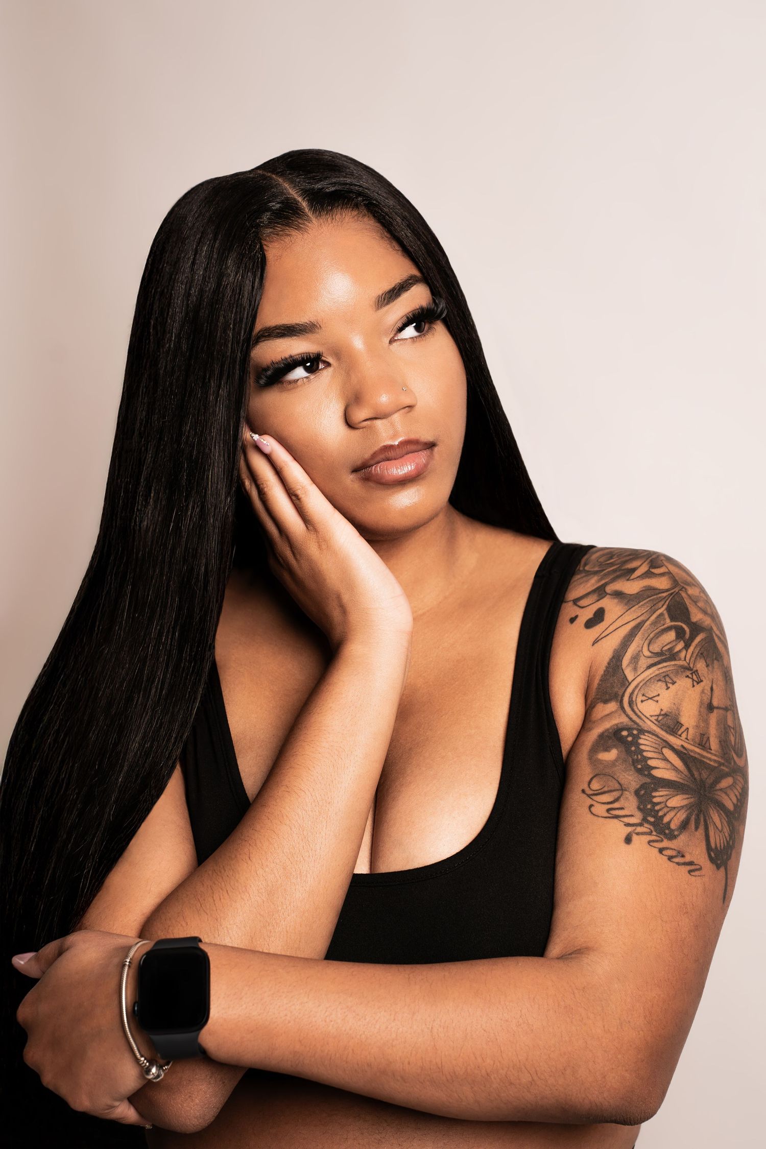 Woman with long black hair and arm tattoos posing thoughtfully with hand on cheek wearing a black top and smartwatch.