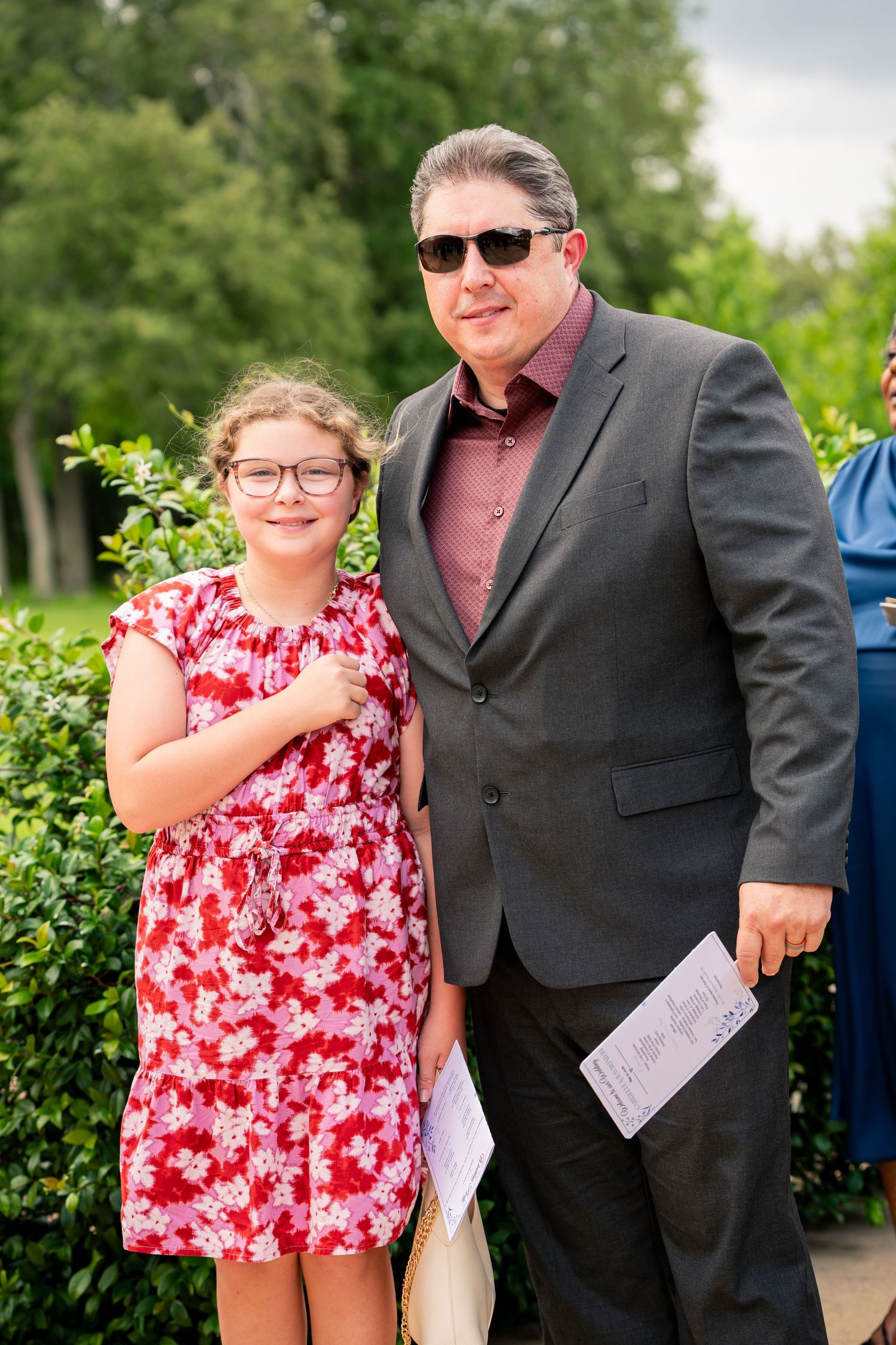 Man in gray suit and sunglasses poses with young girl in floral dress outdoors.
