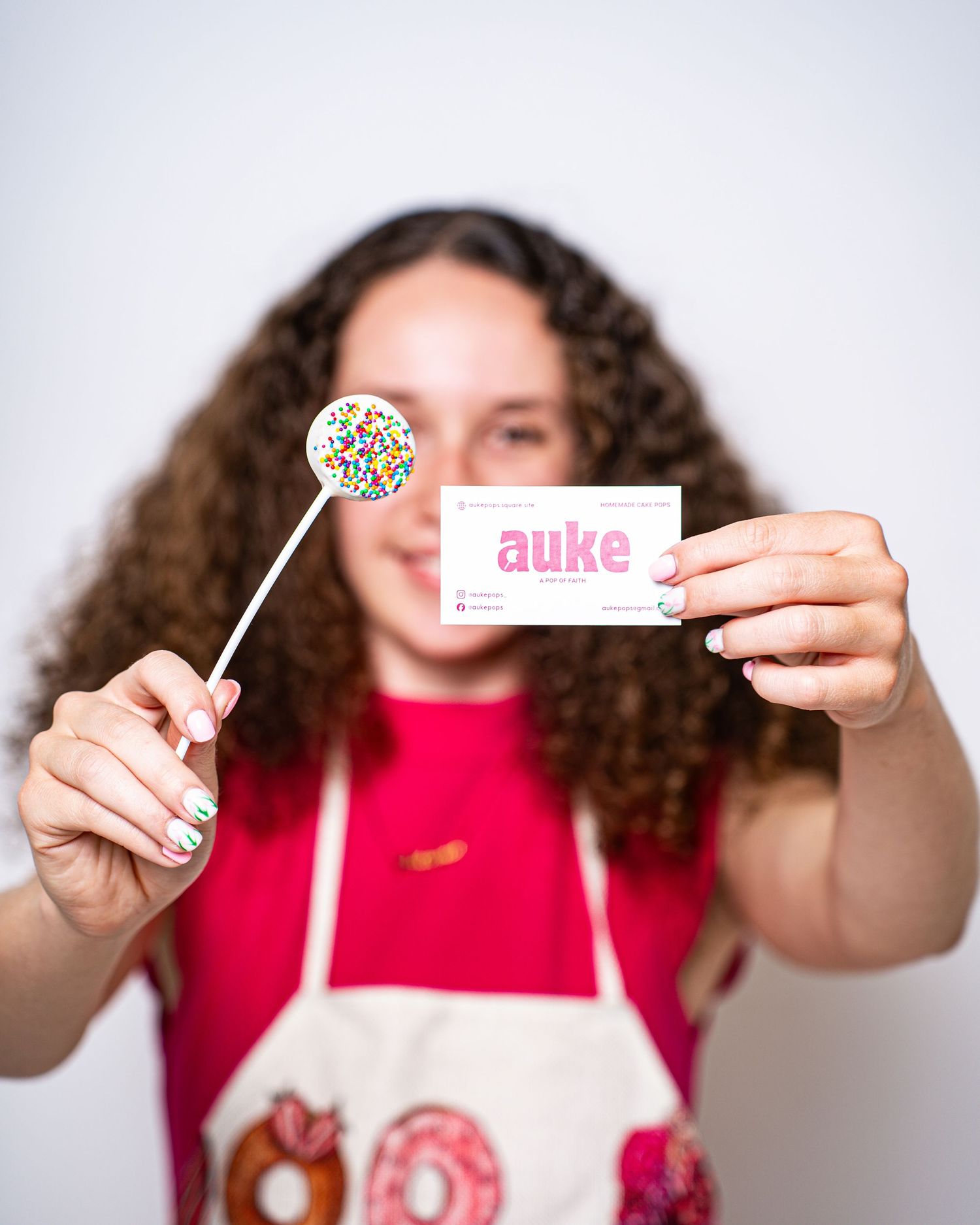 Young woman in baking apron extending a sprinkle lollipop and Auke business card toward the camera.