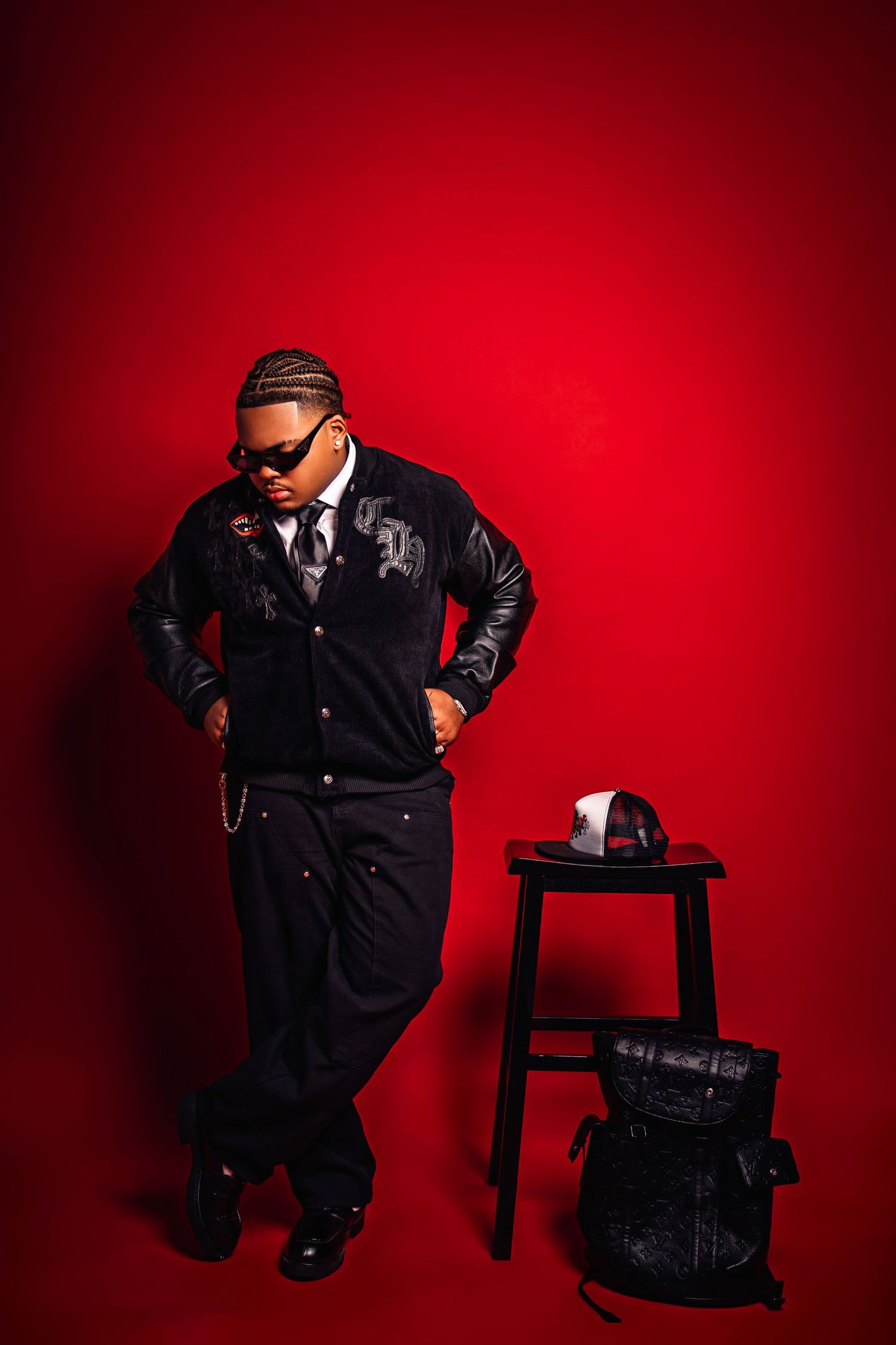 Stylish person in black varsity jacket and sunglasses posing against bold red studio background.