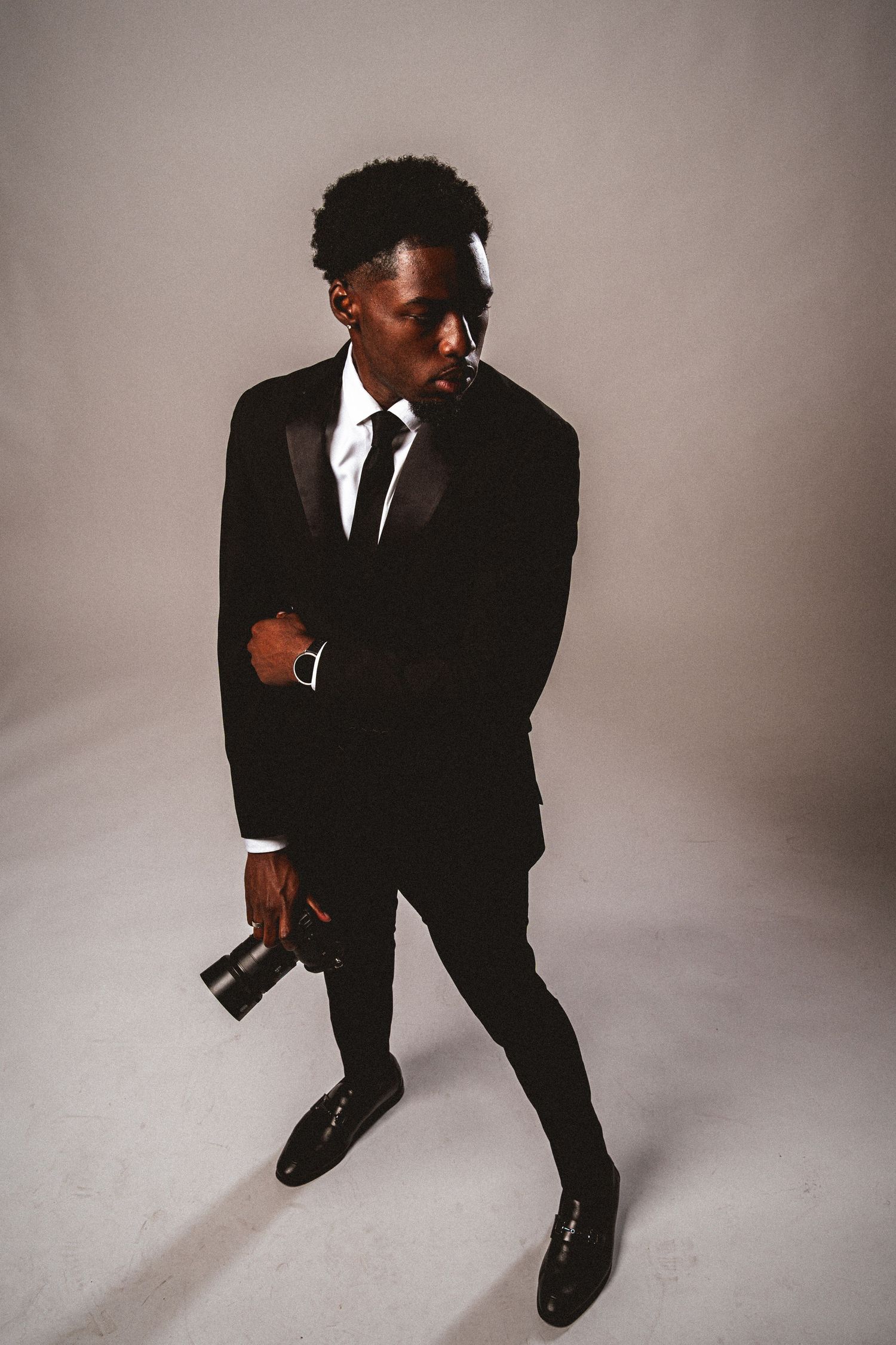 Young Black man in black tuxedo holding a camera while looking sideways on a gray background.