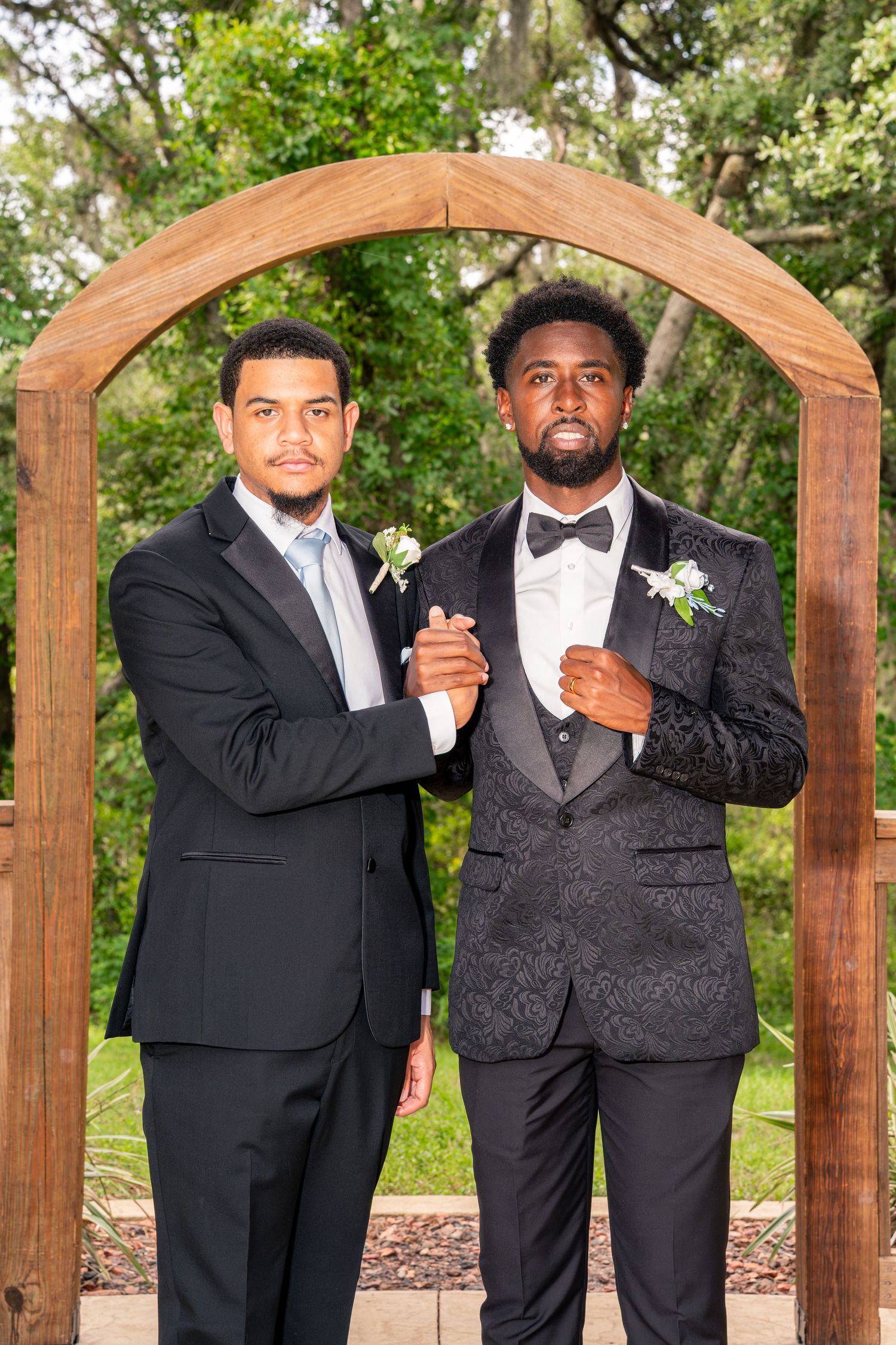 Two groomsmen in black tuxedos pose confidently before a wooden arch at an outdoor wedding ceremony.