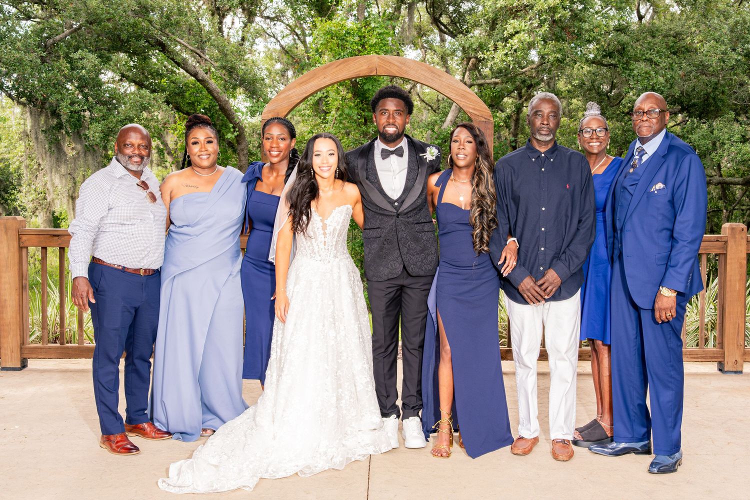 Wedding party group photo with bride and groom under wooden arch outdoors.