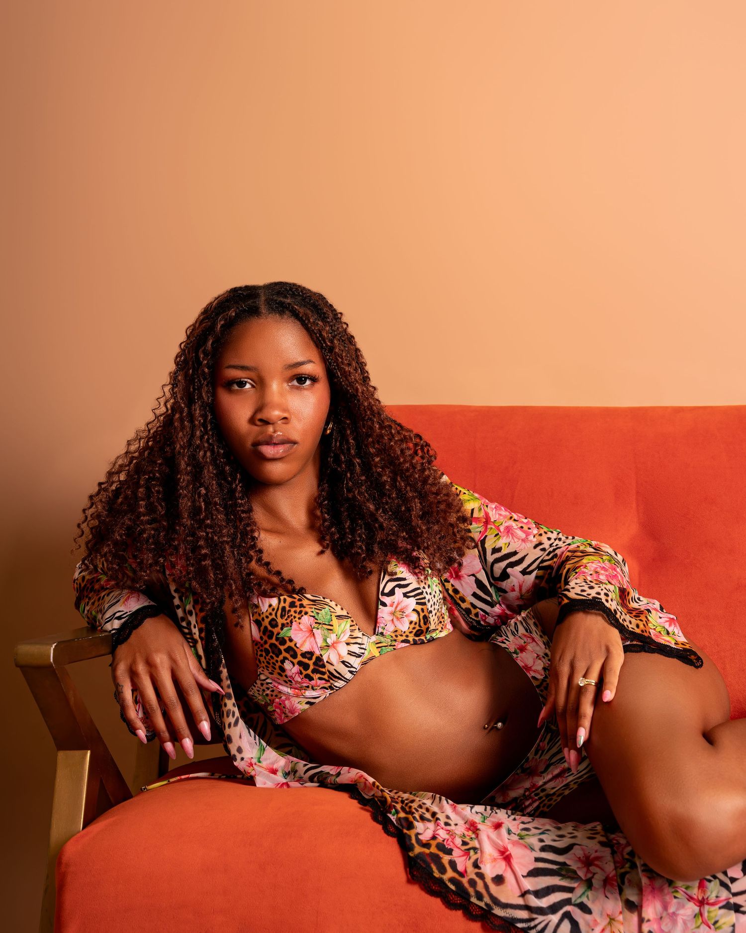 Young woman in floral print bikini set poses on orange velvet chair against warm terracotta background.