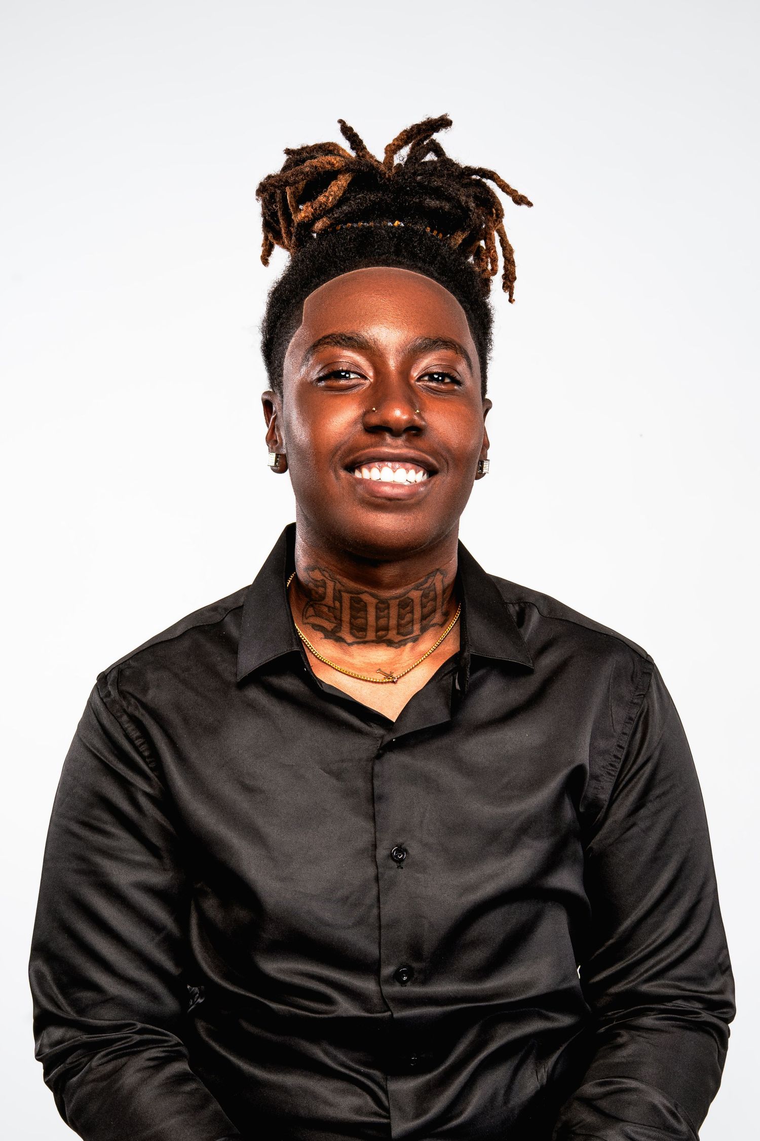 Smiling young Black man with dreadlocks and neck tattoo wearing a black satin shirt against white background.