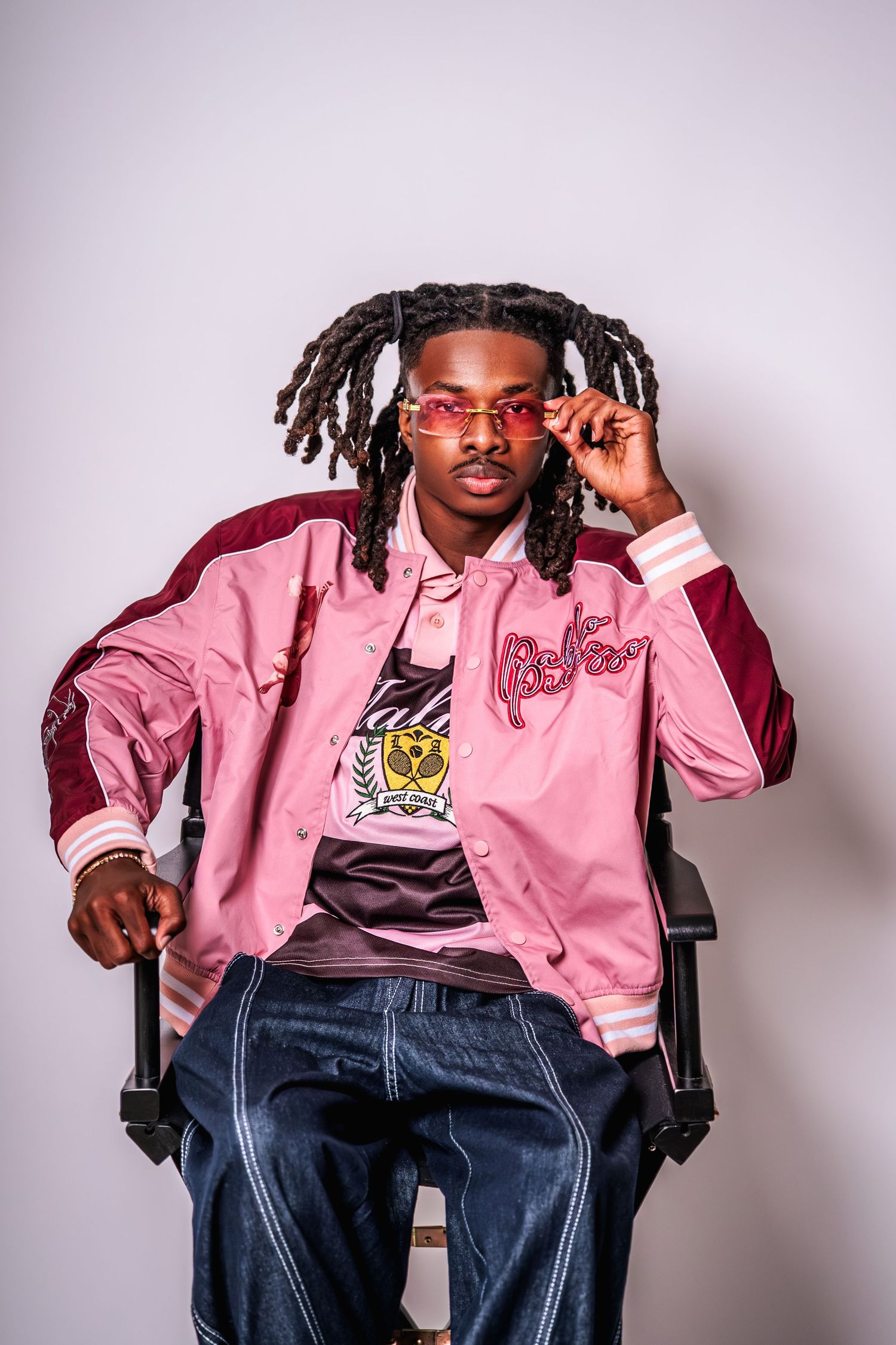Young man with dreadlocks wearing pink sunglasses and a pink varsity jacket seated confidently in a directors chair.