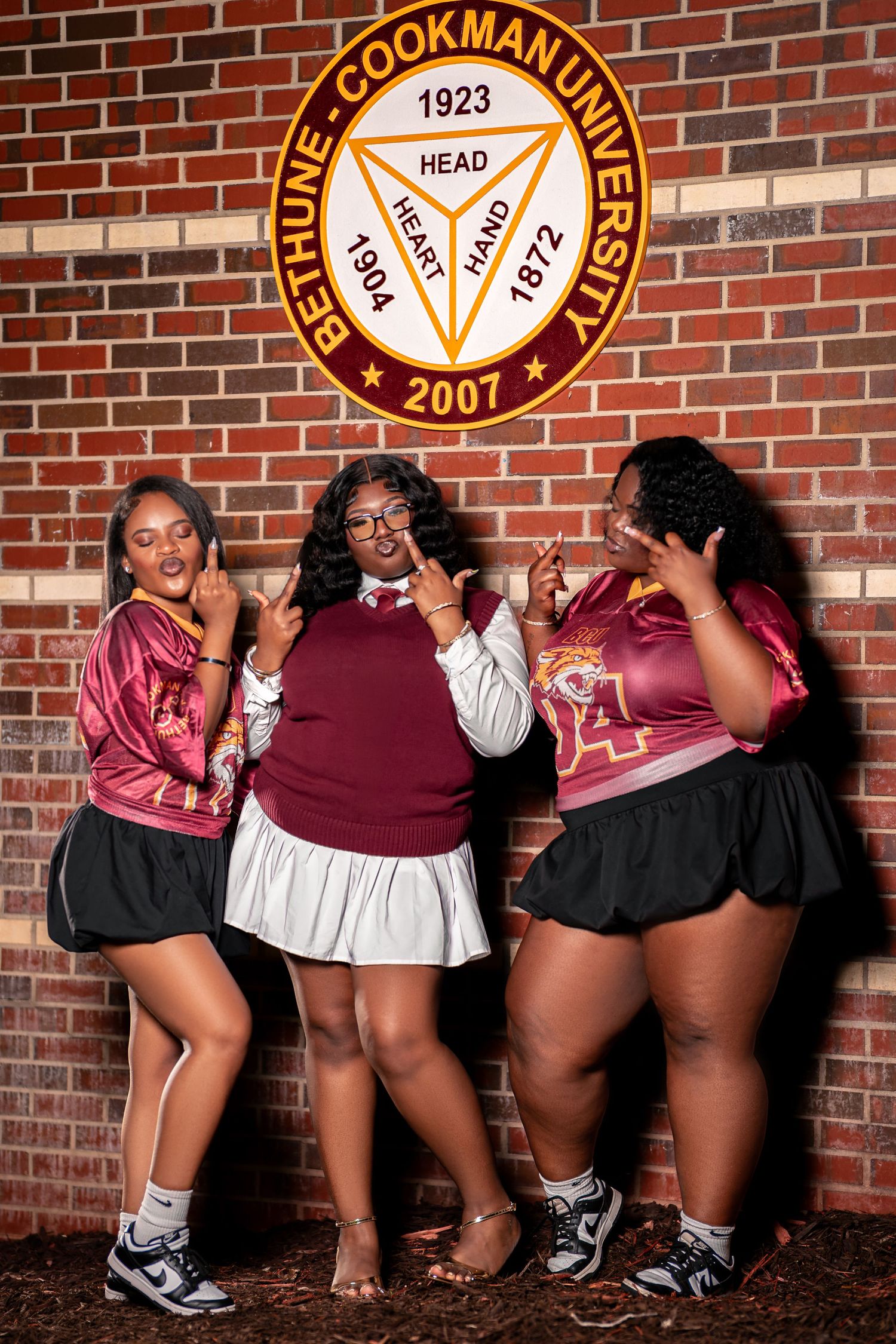 Three young Black women strike fun poses beneath Bethune-Cookman University seal on a brick wall.