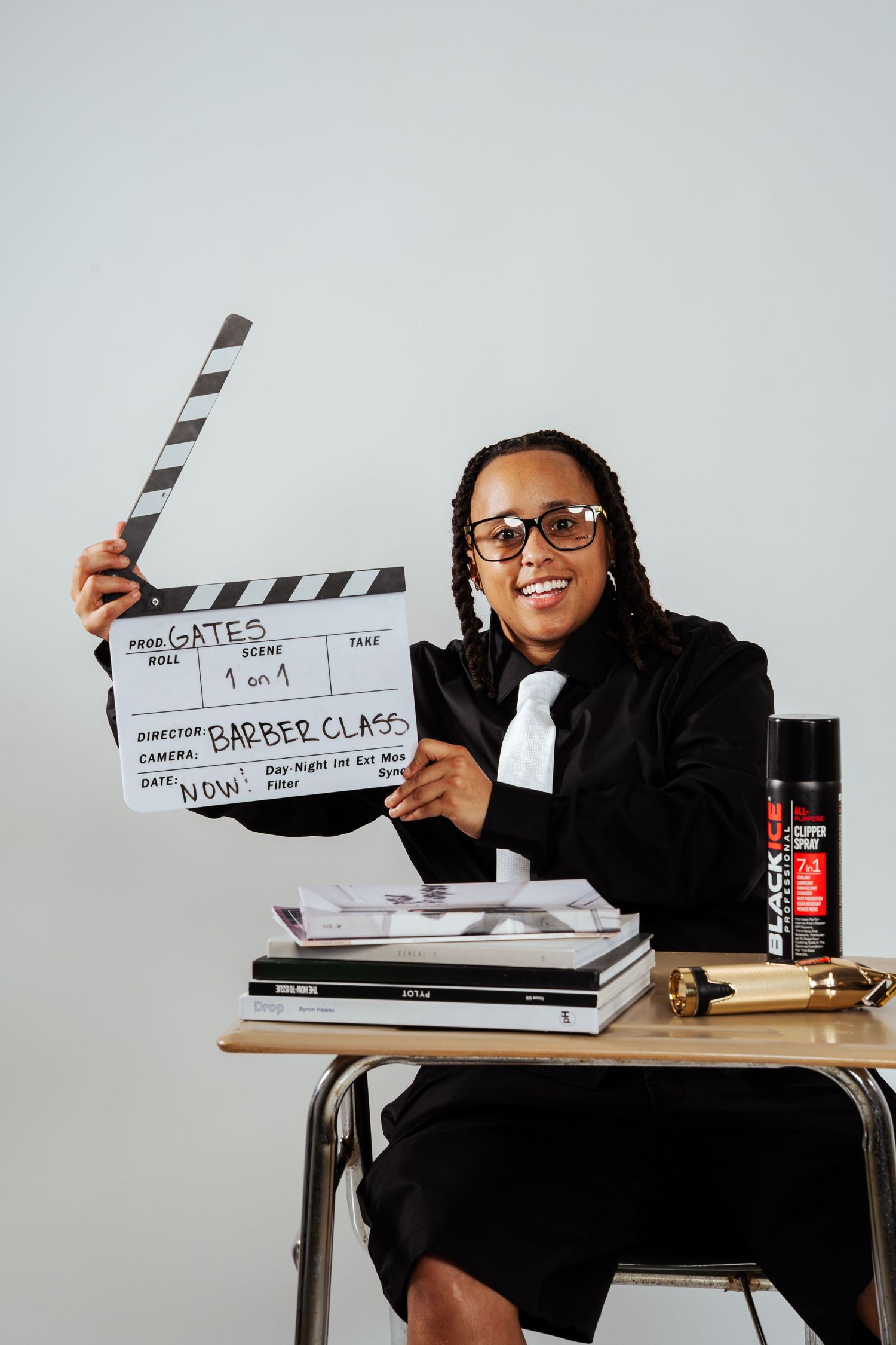 Person in black suit smiling while holding Gates Barber Class film clapperboard at desk.
