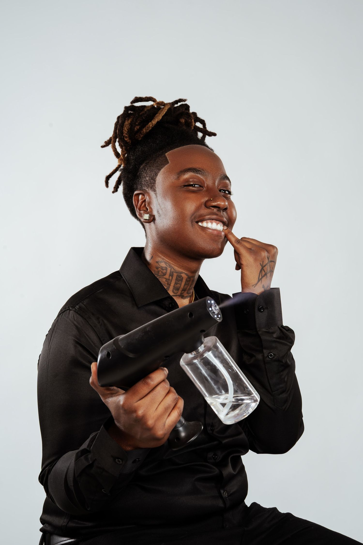 Smiling young Black man with dreadlocks holding a glass bong device while posing in a black satin shirt.