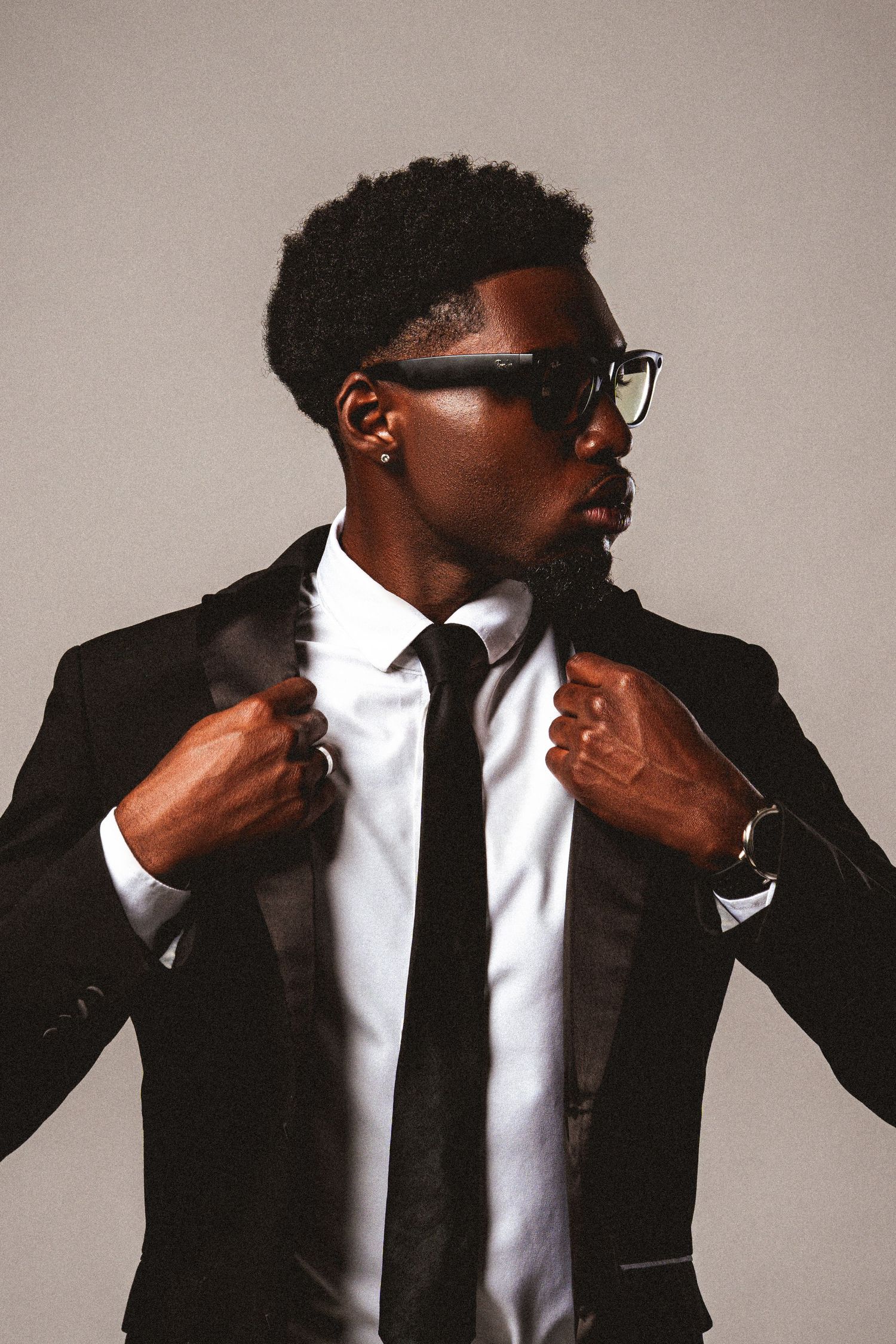Black man in tuxedo adjusting jacket lapels wearing glasses, side glance.