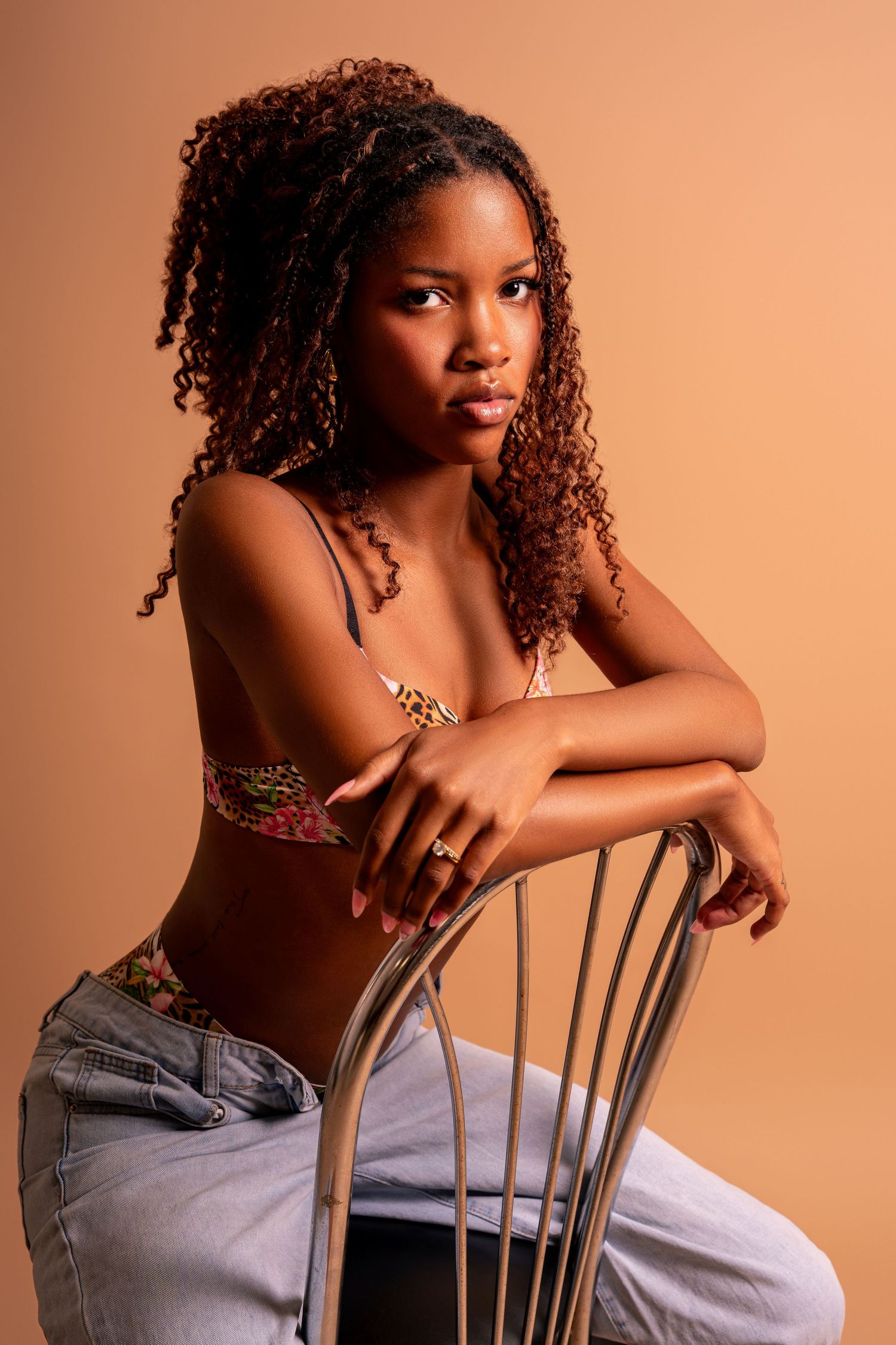 Young woman with curly hair poses on a metal chair wearing a floral bralette and jeans on a beige background.