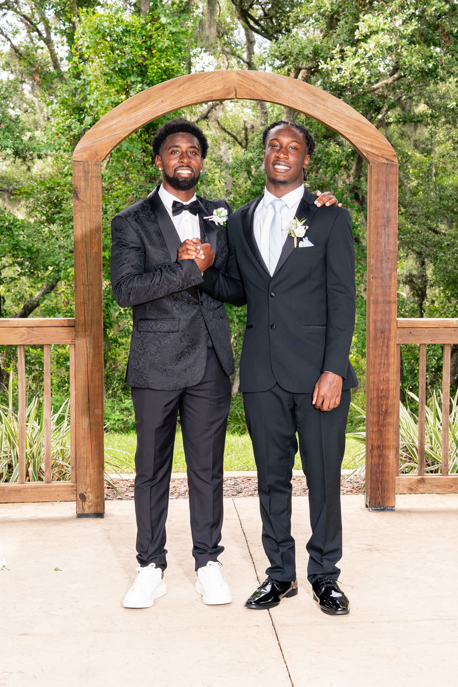 Two well-dressed men in black tuxedos posing together under a wooden arch at an outdoor wedding venue.
