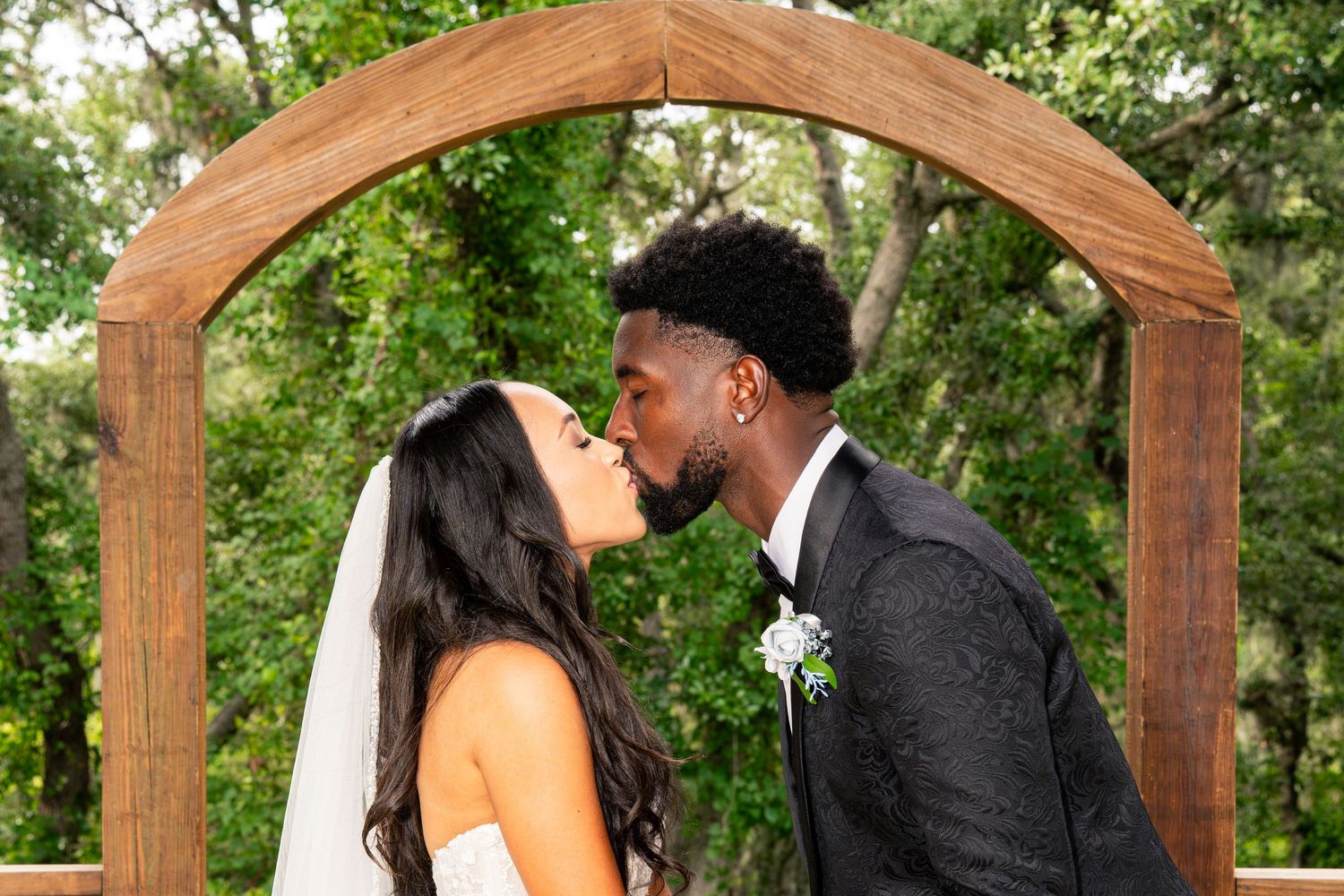 Bride and groom sharing first kiss under rustic wooden arch at outdoor wedding.