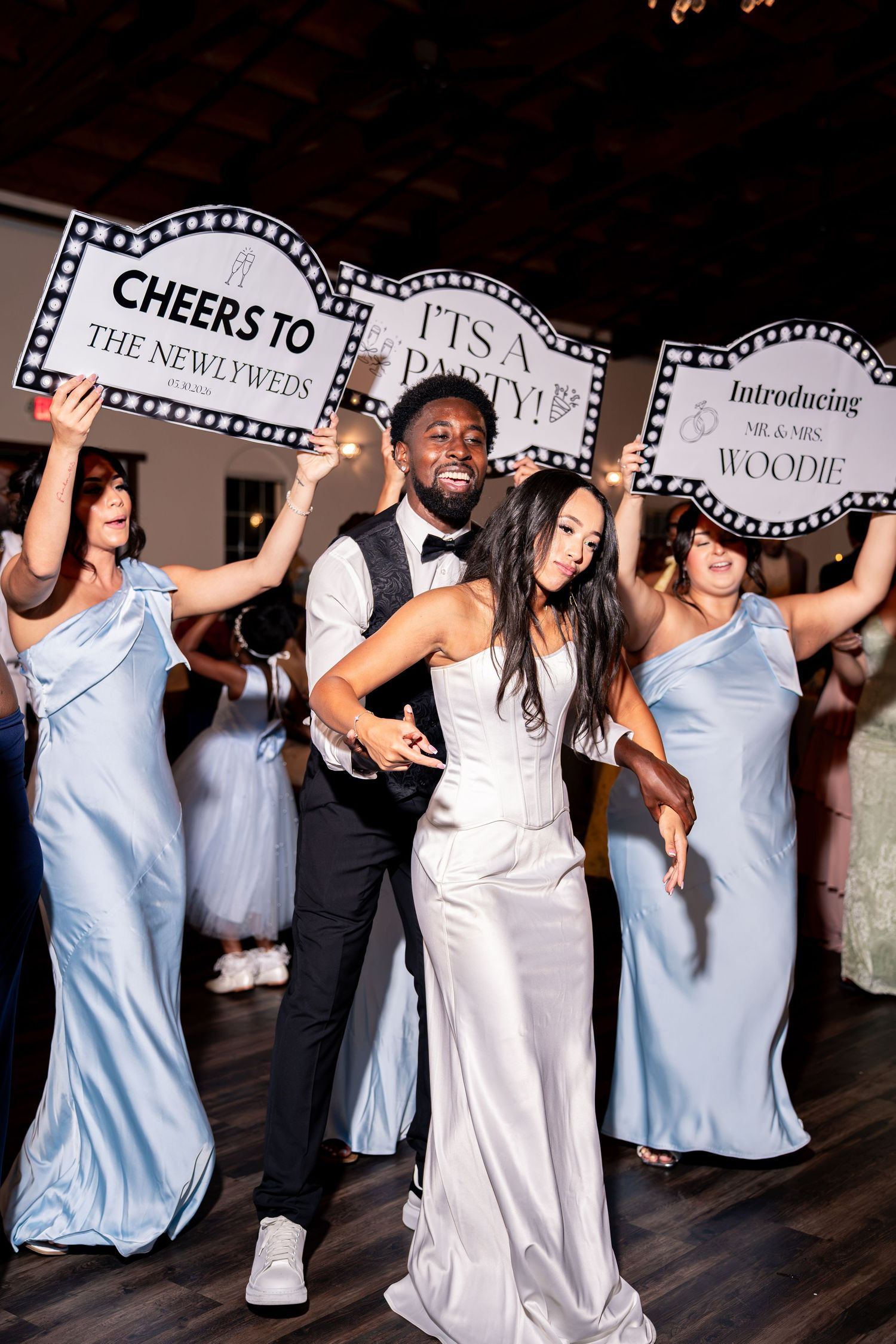 Newlyweds dance joyfully as bridesmaids hold marquee signs reading Cheers to the Newlyweds and Introducing Mr and Mrs Woodie.