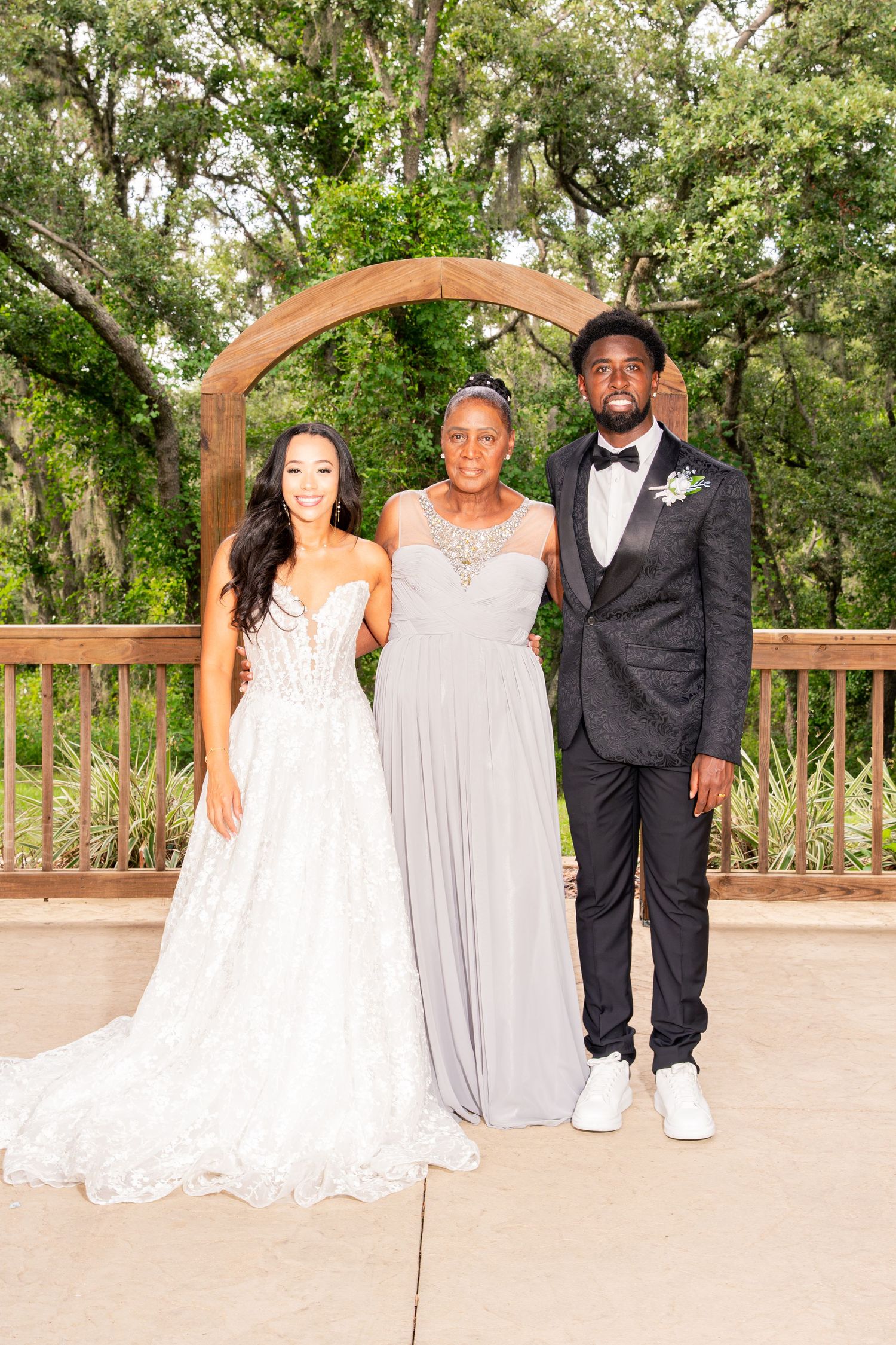 Bride and groom pose with older woman in gray gown under wooden arch outdoors.