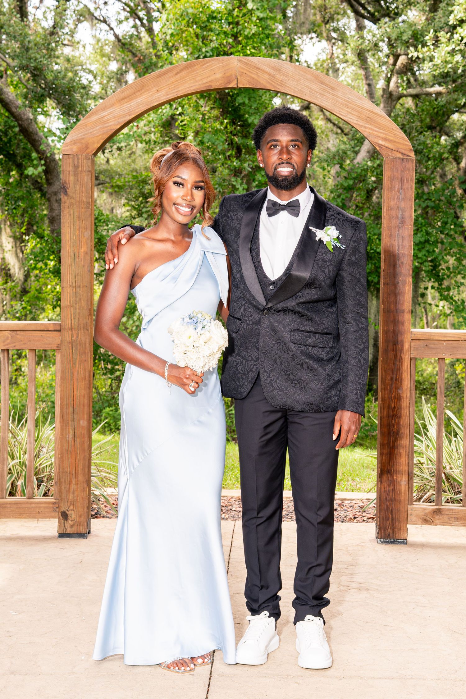 Couple posing under wooden arch, woman in blue gown holding bouquet, man in black tuxedo.