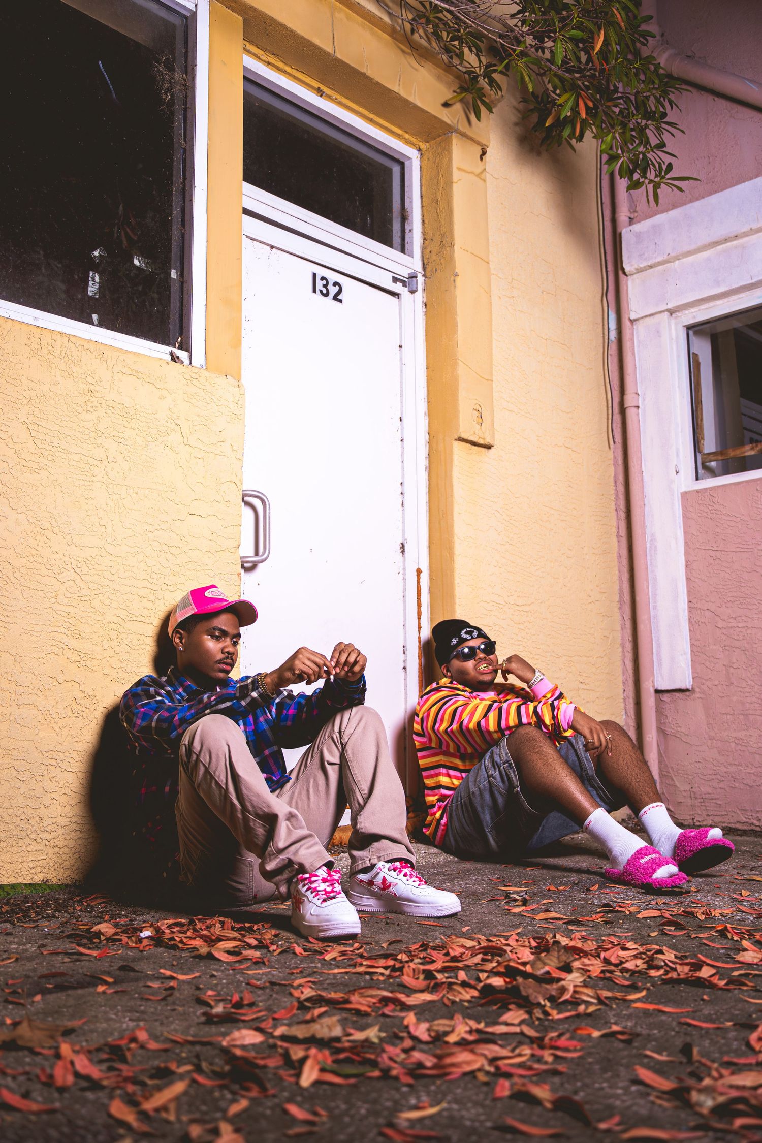 Two young men sitting on ground against yellow building surrounded by fallen leaves wearing vibrant streetwear outfits.