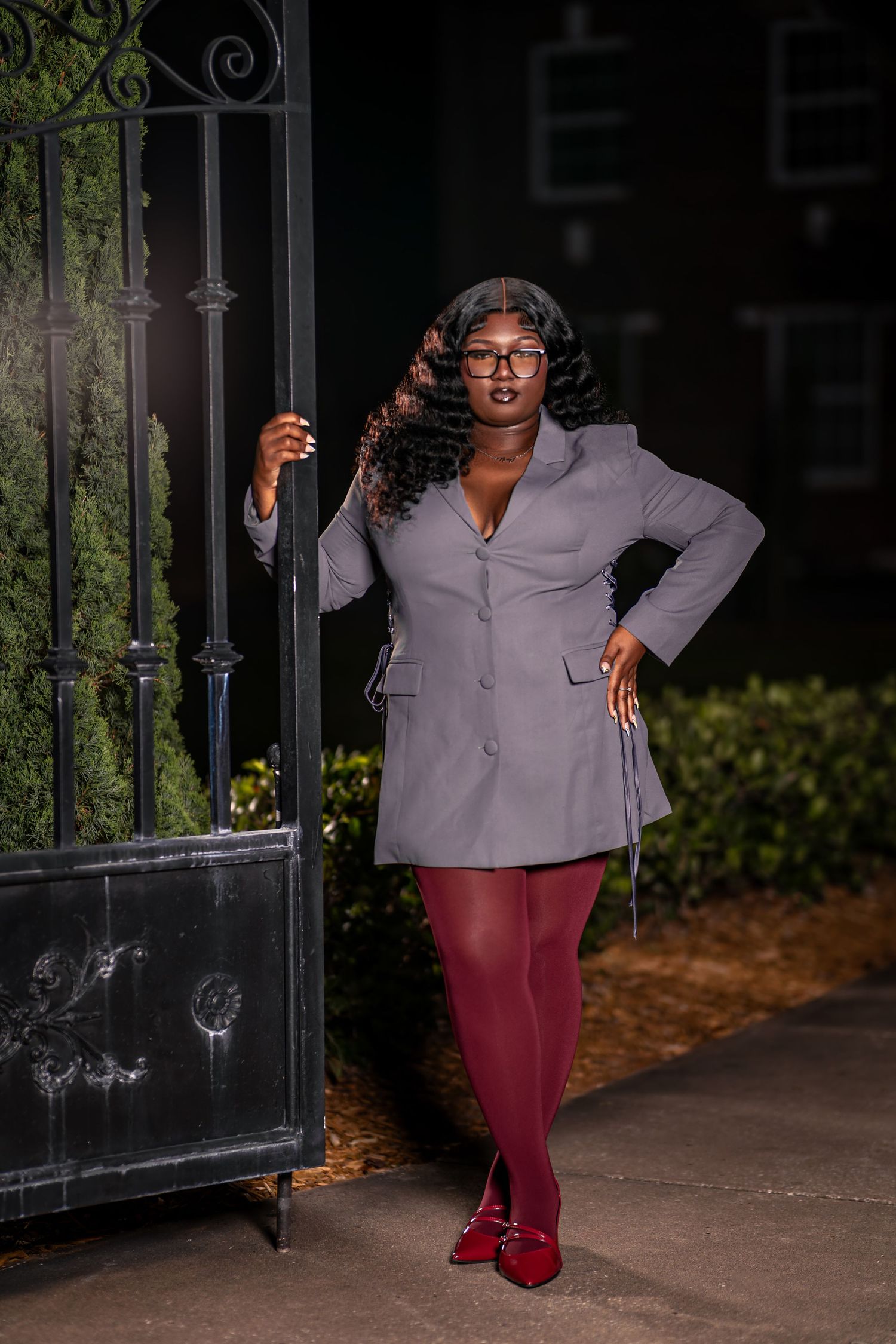 An elegant Black woman in gray blazer dress and burgundy tights poses by ornate iron gate at night.