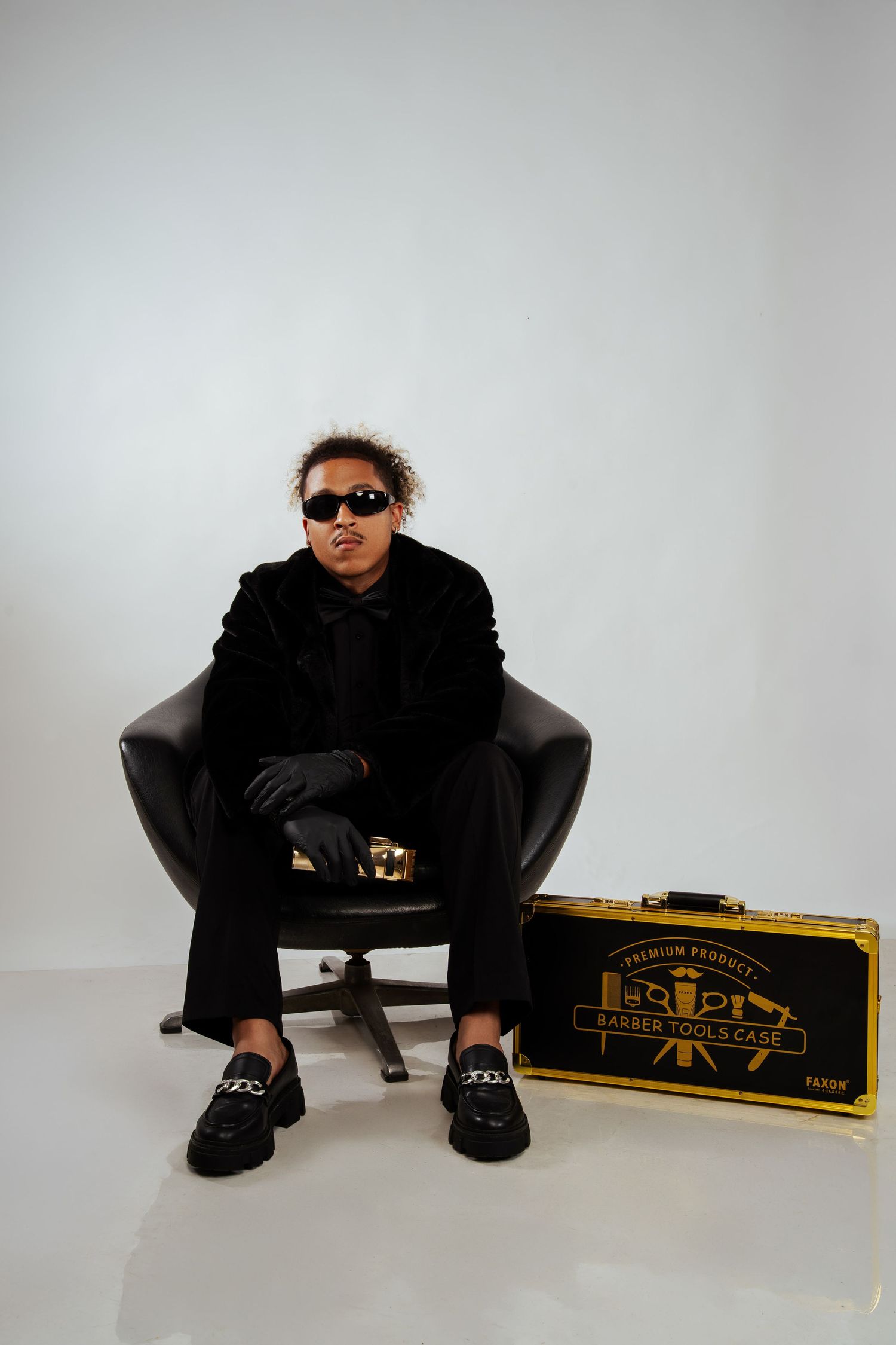 Confident person in black fur coat sitting in leather chair beside gold barber tools case.