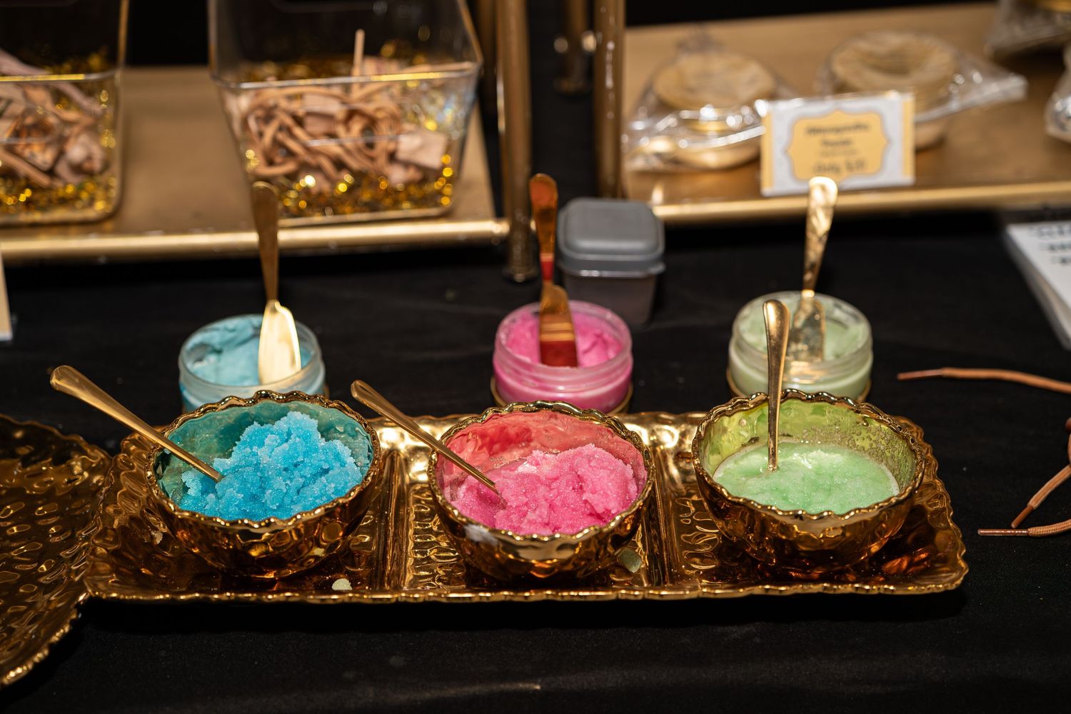 Colorful sugar scrubs displayed in gold bowls on black tablecloth at vendor booth.