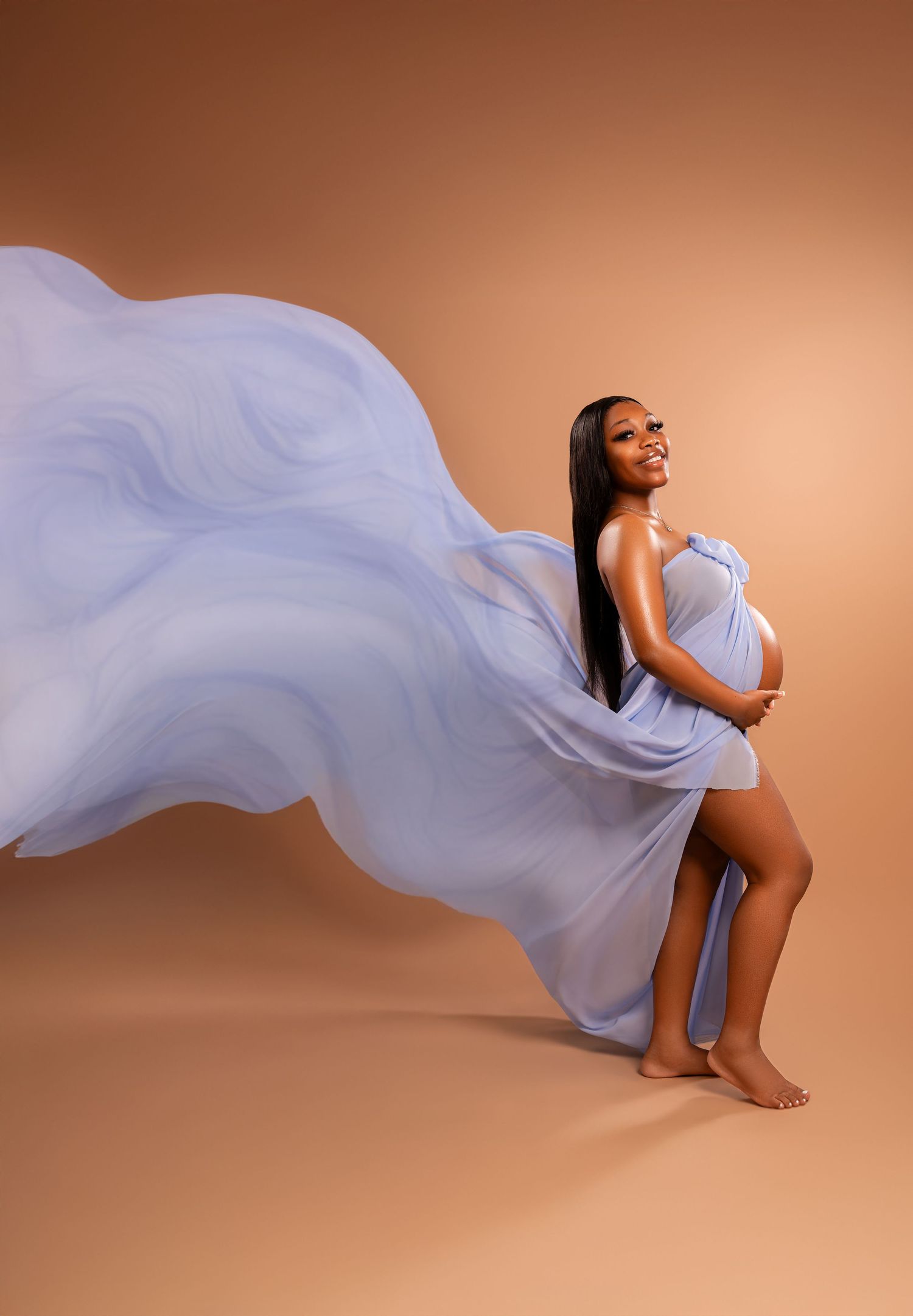 Pregnant woman in flowing blue chiffon fabric poses gracefully against warm beige background in elegant maternity portrait.