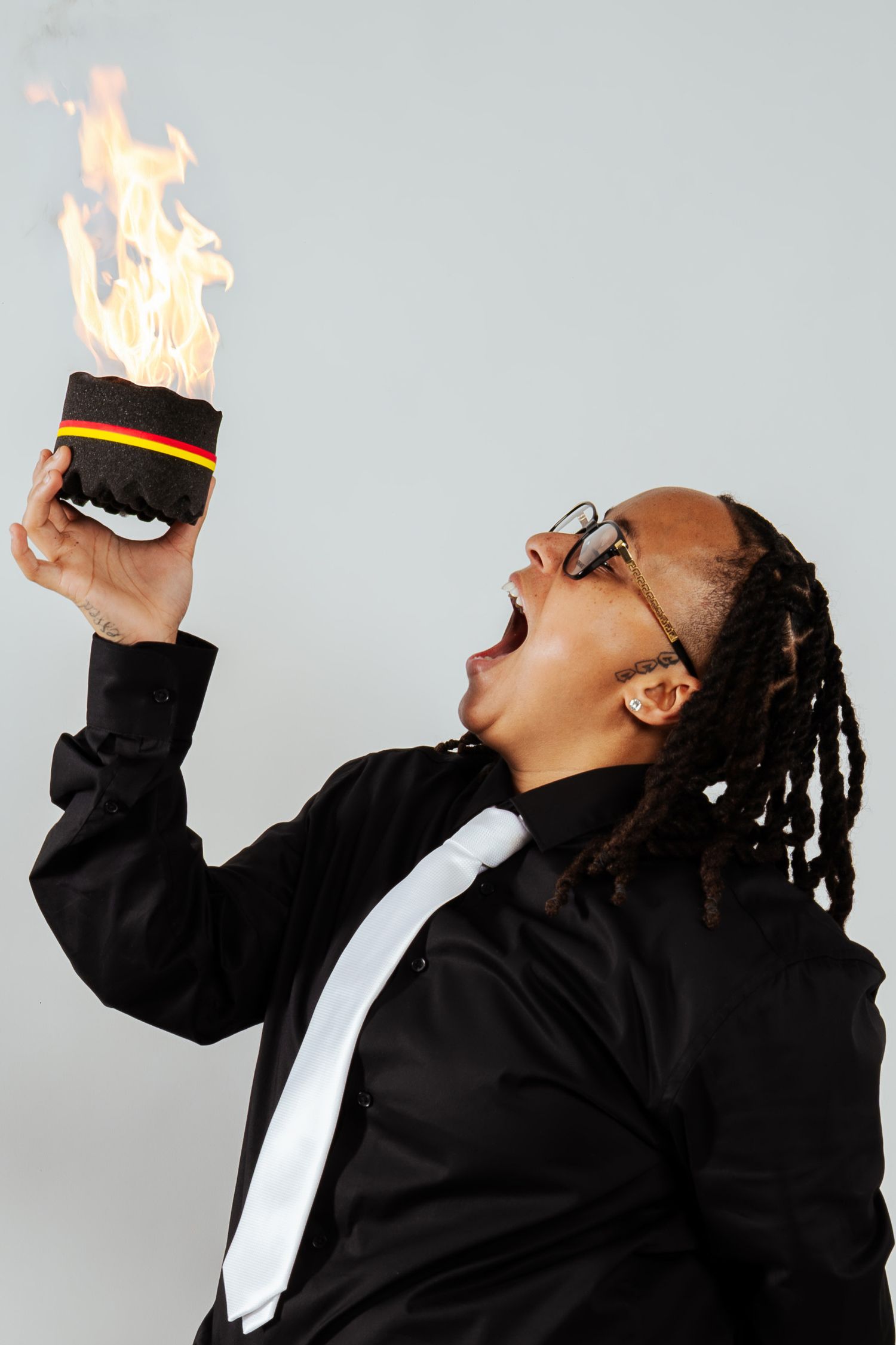 Person in black shirt holding flaming object with mouth open wide against gray background.