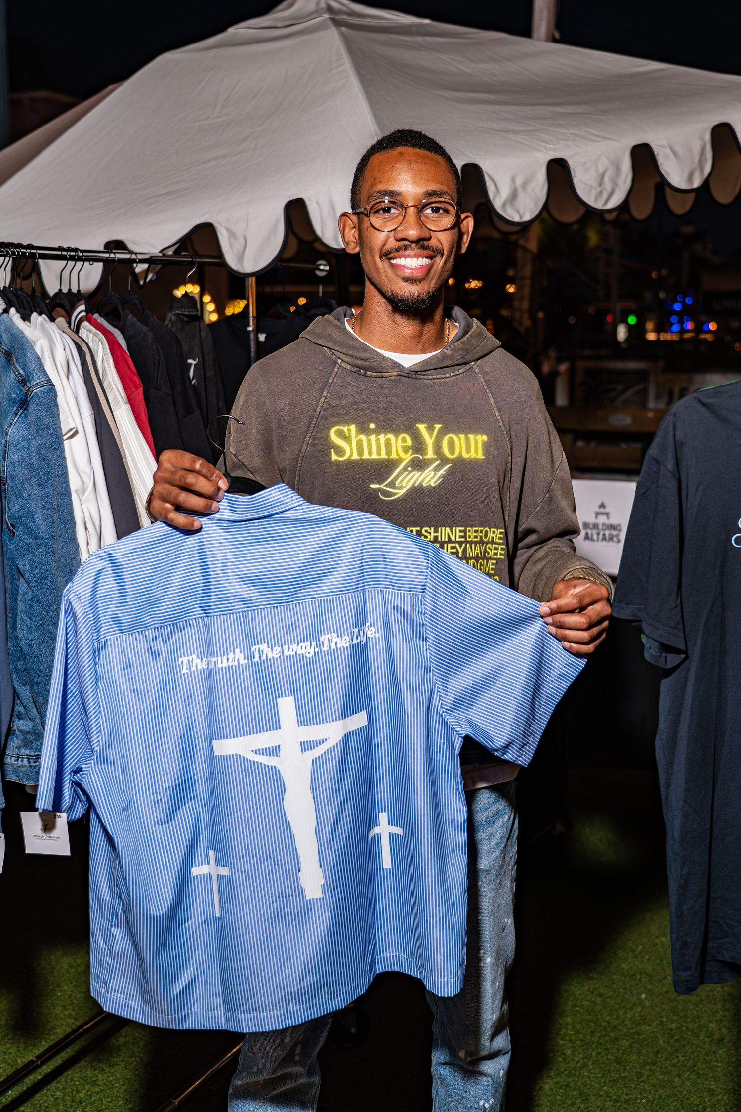 Smiling man holding up a blue Jesus cross shirt at Through Christ Brand outdoor pop-up.